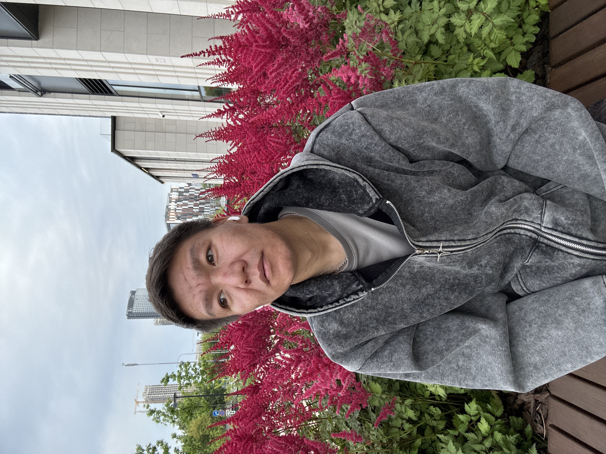 A medium shot captures a man sitting on a wooden bench in front of a vibrant display of bright pink Astilbe flowers. The man, with short, dark hair and light skin, wears a grey zip-up hoodie over a grey t-shirt and a silver necklace. He has white earbuds in his ears and looks off to his right. Behind the flowers, modern buildings with clean lines and glass windows dominate the background, suggesting an urban setting. The sky is overcast with grey clouds. To the left, construction cranes and tall buildings can be seen, indicating ongoing development within the city of Khimki, Russia. The overall scene is one of a man taking a moment of repose amidst a cultivated urban landscape.
