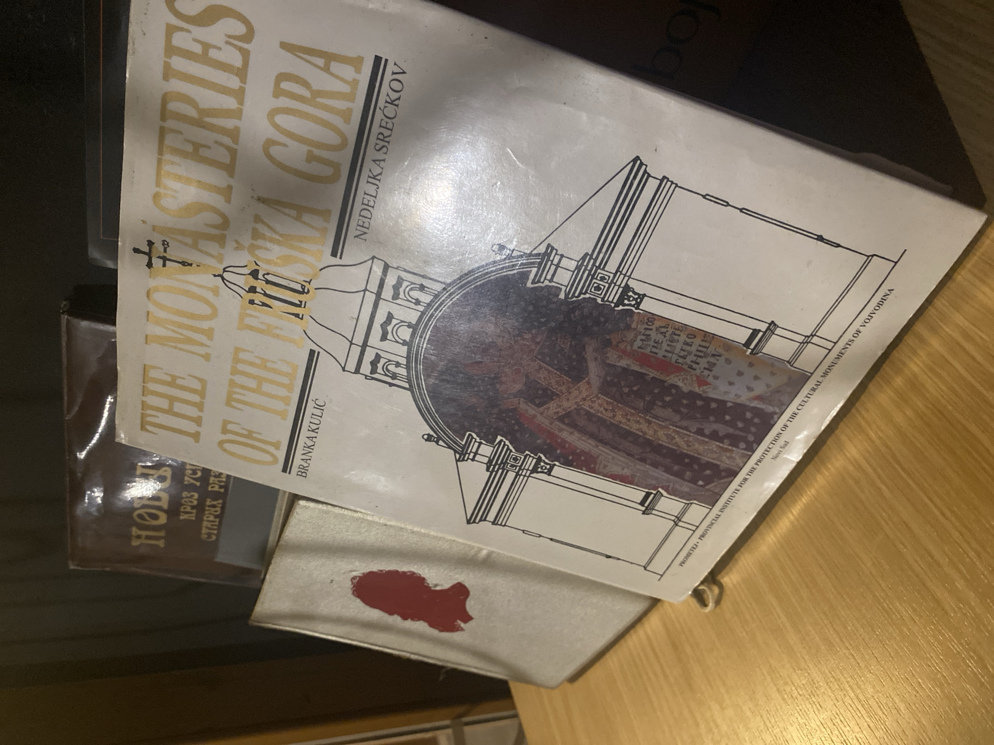 The image shows a book titled "The Monasteries of the Fruška Gora" by Branka Kulić and Nedeljka Srećkov, published by PROMETEJ - PROVINCIAL INSTITUTE FOR THE PROTECTION OF THE CULTURAL MONUMENTS OF VOJVODINA, Novi Sad. The book has a white cover with gold lettering and a drawing of a monastery. There is another book partially visible behind it, with a brown cover. A red stain is also present on the cover of the book about monasteries. The background is a wooden surface.