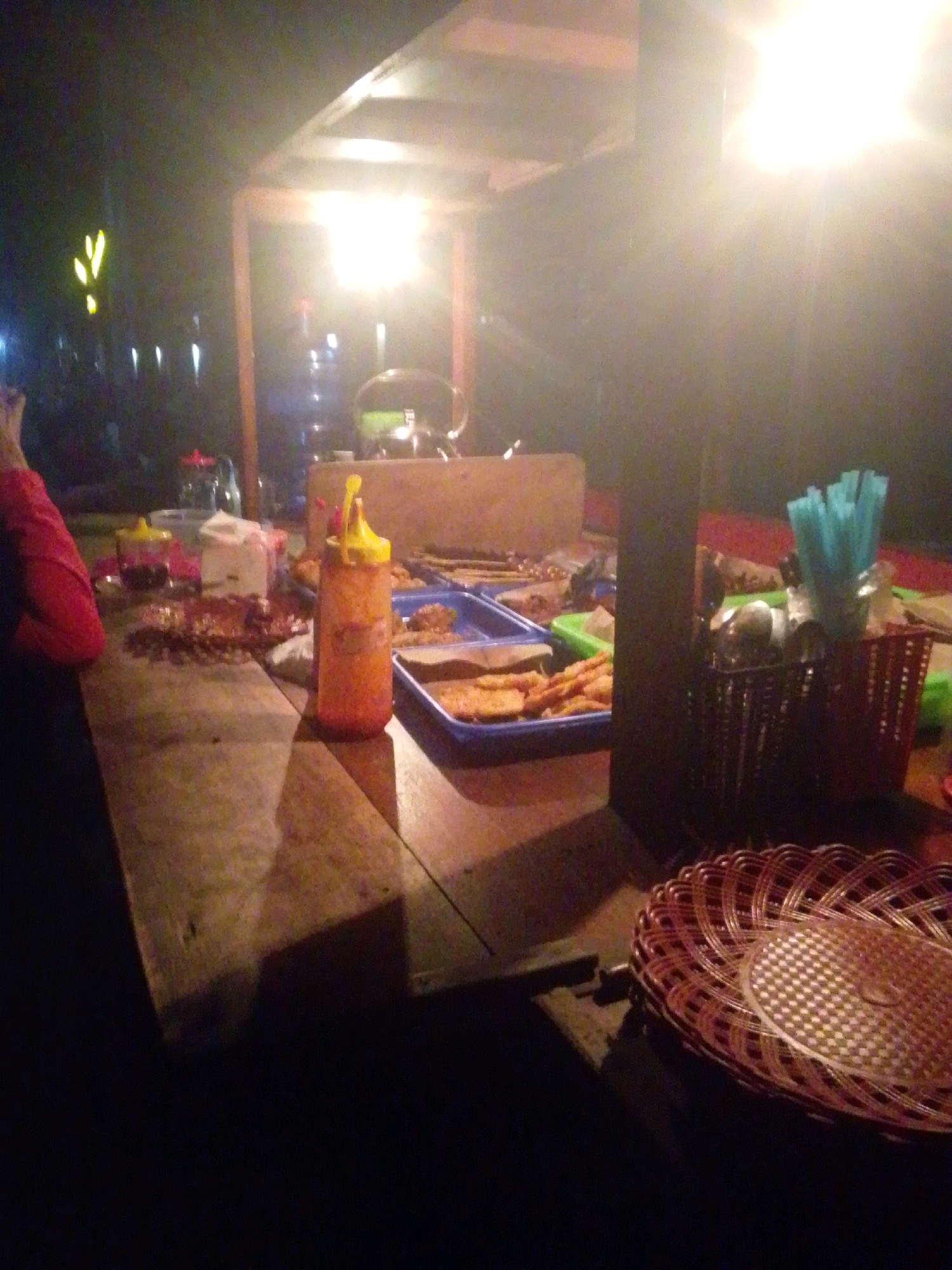 The image depicts an outdoor food stall at night, likely in Bedono Kluwung, Indonesia. The scene is illuminated by a bright overhead light, casting a warm glow on the wooden table and various food items. In the foreground, a wooden table is laden with plates of food, including what appears to be fried snacks and some sort of relish in a squeeze bottle. To the right, a basket holds blue plastic straws and other utensils. In the background, a person wearing a red garment is partially visible, suggesting they are either a customer or staff. The stall seems to be set up under a simple wooden canopy.