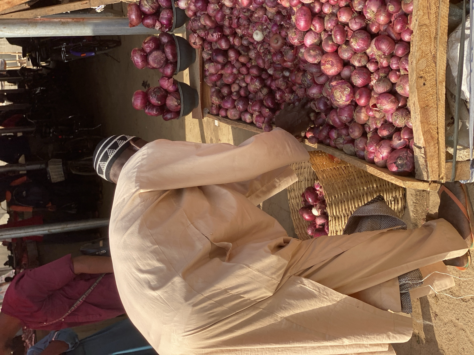 An outdoor market scene in Kafanchan B, Nigeria, unfolds under bright daylight. The primary subject is a man, seen from behind, wearing a light tan or beige traditional long-sleeved garment and a distinctive black and white patterned Kufi cap. He is positioned beside a large, overflowing display of red onions.
The onions are the central focus of the stall, piled high on a wooden structure, possibly a table or cart. Additional onions are visible in several dark-colored buckets and a woven basket placed on the ground. The man's right hand is reaching into the large pile of onions, suggesting he is either arranging them, selecting them, or preparing a portion for sale.
In the background, on the left side of the frame, several bicycles are parked or stacked, indicating a bustling market environment. A partial view of another person, dressed in a purple garment, is visible in the lower left corner. The ground appears to be dusty earth or concrete. The strong illumination suggests a sunny day. No discernible text is visible in the image.