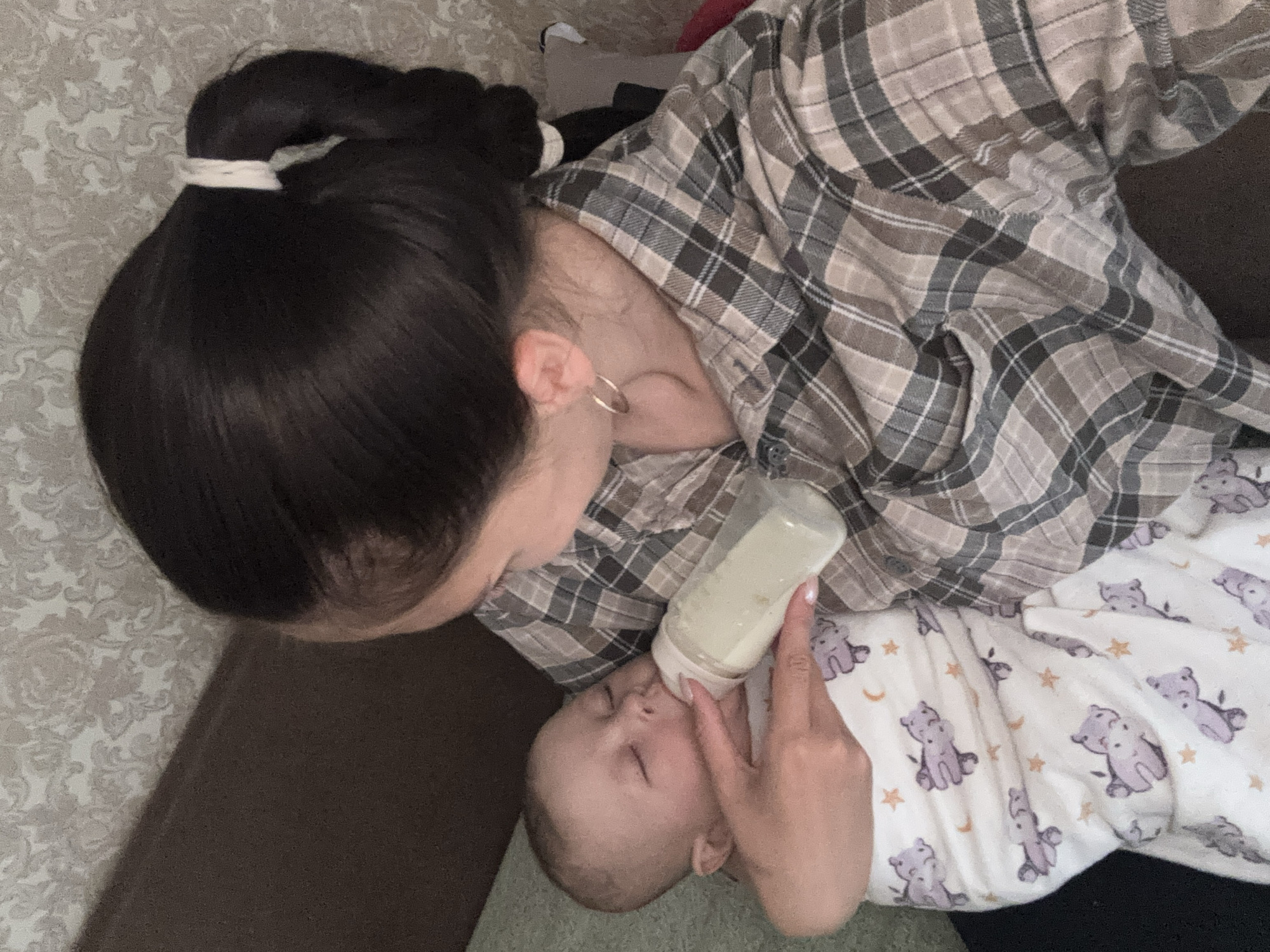 A woman is feeding a baby. The woman has dark hair pulled back in a ponytail with a white tie. She is wearing a gray and tan plaid shirt. The baby is lying down, wearing a white sleeper with gray teddy bears and orange stars and moons. The woman is holding a bottle to the baby's mouth. The location is not evident in the image, so I cannot include Sochi, Russia.