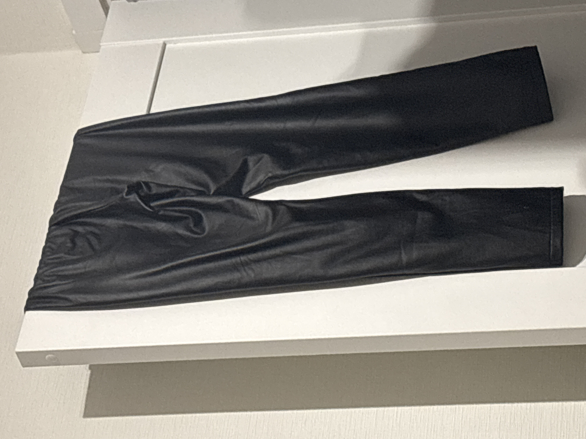The image shows a pair of black, shiny leggings resting on a white shelf or surface. The leggings are wrinkled and appear to be made of a material that reflects light. The shelf is simple and appears to be mounted on a wall or inside a closet. The environment is indoors and the lighting is neutral. It is impossible to provide a location in the city of Vsevolozhsk, Russia, with only this image.