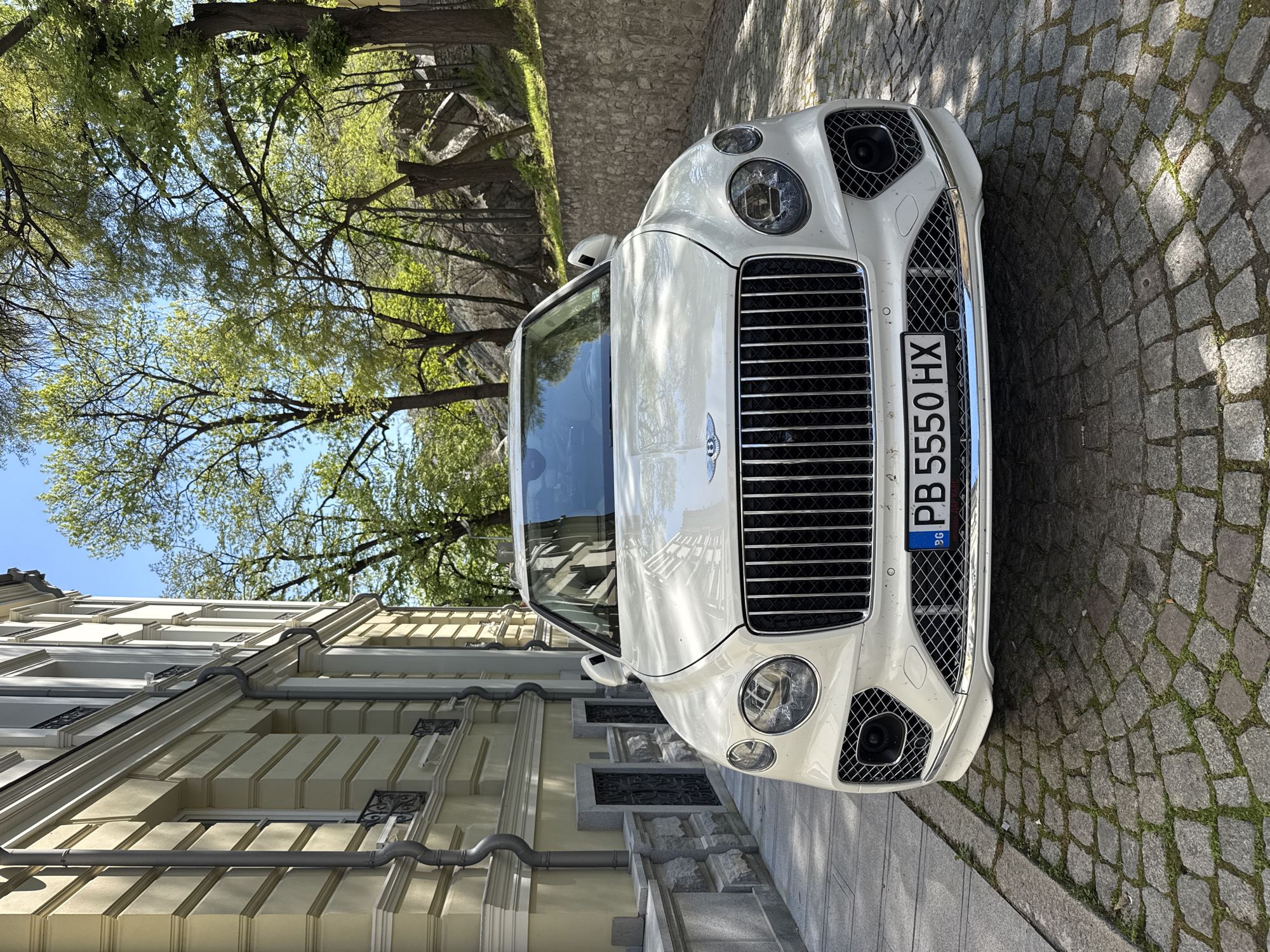 The image shows a white Bentley Continental parked on a cobblestone street next to a light-yellow building with decorative window surrounds and rain gutters. The car's license plate reads "PB 5550 HX." In the background, there are trees with fresh spring leaves and a stone retaining wall. The scene appears to be in a well-maintained, possibly historic area. It is difficult to determine the exact location within Plovdiv without further information, but the architecture suggests a central or older district. There are no people visible in the image. The subject is clearly the Bentley, suggesting wealth and luxury.