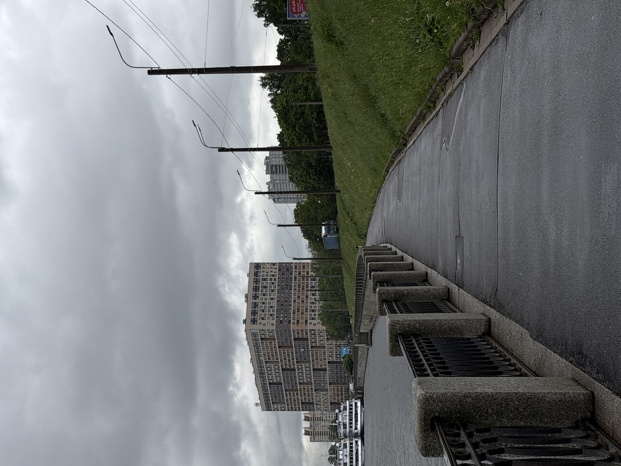 The image depicts a waterfront scene in Saint Petersburg, Russia, under an overcast sky. A pedestrian walkway runs along a river or canal, separated from the water by a low concrete barrier with metal grates. A grassy slope rises on the other side of the walkway, lined with trees and lampposts. In the background, a tall, multi-story building with a patterned facade is visible, along with other buildings and structures. Several boats are moored near the walkway. The scene appears to be urban, possibly a river embankment in Saint Petersburg.