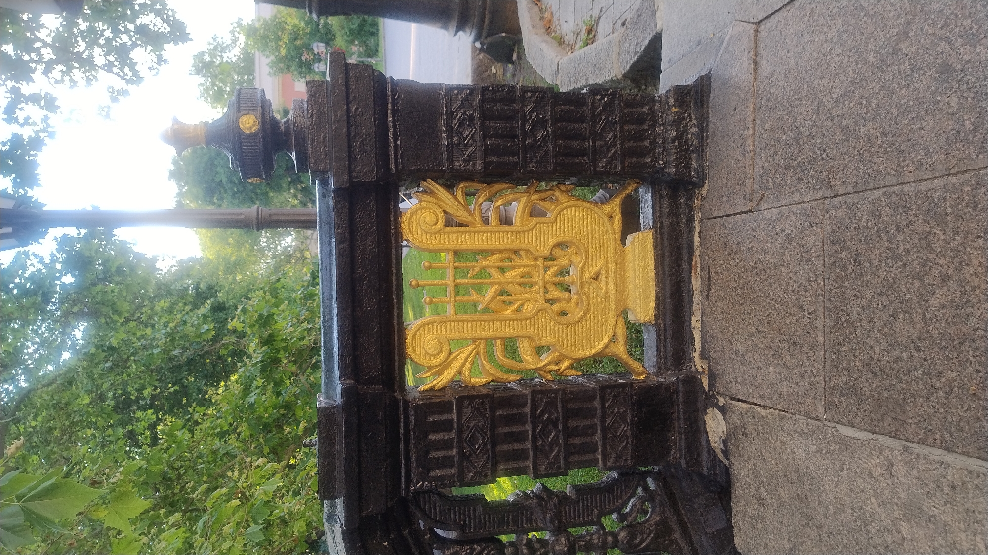 This is a close-up, vertically oriented, eye-level photograph of a decorative black metal fence post. The fence post has a golden lyre emblem in the center, with ornate scrollwork above and below it. The fence post itself is elaborately carved with a repeating geometric pattern. To the left of the fence post is a lush green canopy of trees and foliage, and a lamppost can be seen in the upper left quadrant of the image. The background to the right of the fence post consists of a grey, speckled granite wall. The lighting suggests it is daytime, possibly late afternoon, with sunlight filtering through the leaves and creating a bright glare on the lamppost. The scene appears to be outdoors in a park-like setting or along a decorative pathway. There is no visible text in the image.
