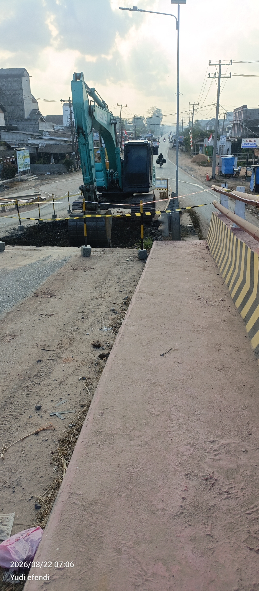 The image captures a scene in Rawajitu Selatan, Indonesia, where road construction is underway. A large, teal-colored excavator is positioned over a freshly dug pit in the road. The pit is bordered by yellow and black striped caution tape strung between vertical poles, some of which are anchored by concrete blocks. The ground around the pit is unpaved and appears to be dirt and gravel. In the background, a paved road stretches into the distance, with vehicles, including motorcycles and cars, visible. Buildings line both sides of the road, suggesting a commercial or residential area. Utility poles with electrical wires are also prominent. A modern street light with a solar panel stands near the center of the image. The sky is overcast with some patches of sunlight breaking through. In the foreground, a section of a paved walkway or elevated platform with a reddish-pink hue is visible, marked with diagonal yellow and black stripes on its edge. A pink plastic bag lies on the ground near the bottom left corner of the image, accompanied by a timestamp and name: "2026/08/22 07:06 Yudi efendi".