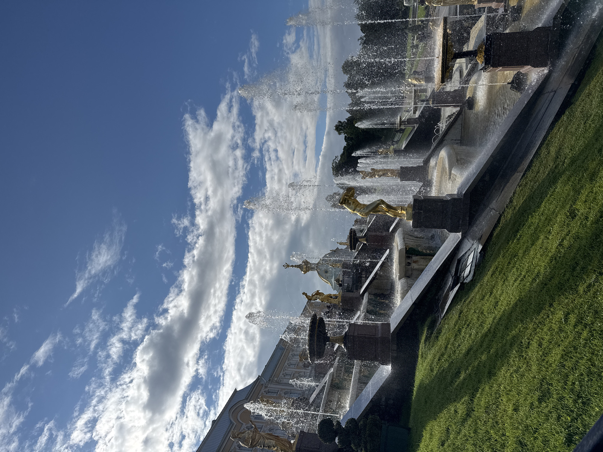 The image depicts the Grand Cascade, a famous fountain complex located in Peterhof Palace, Russia. The scene is dominated by numerous water jets spraying upwards from ornate basins and sculptural elements. Golden statues of mythological figures are positioned throughout the cascade, adding to its grandeur. The water flows down a series of tiered basins and channels, creating a dynamic and impressive spectacle. In the background, a large building with a dome-like structure, likely part of the palace, is visible. The sky above is blue with scattered white clouds, and a lush green lawn slopes down from the left of the frame. The overall impression is one of opulence and natural beauty, characteristic of the Baroque style of Peterhof.