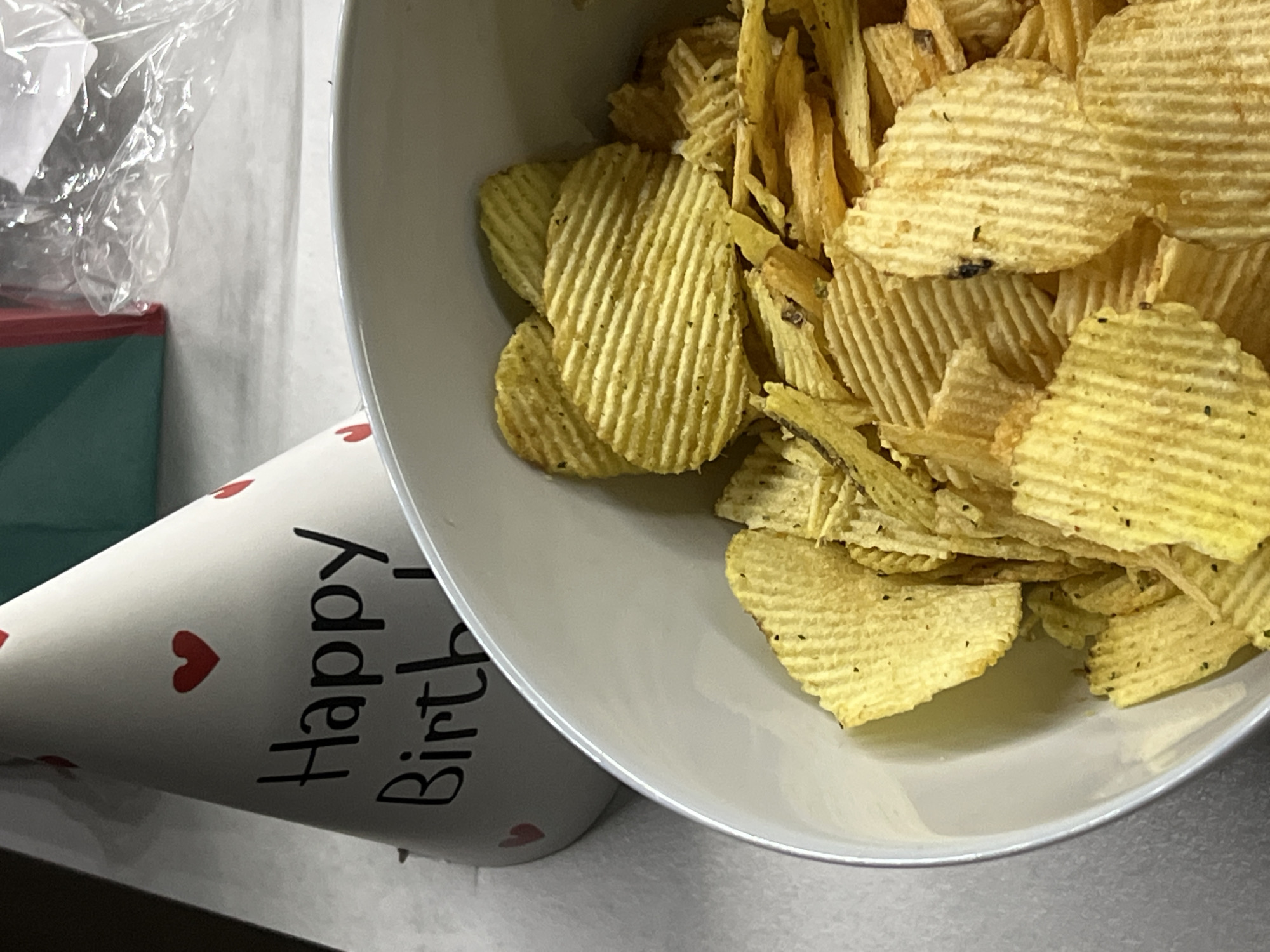 A white, conical party hat, adorned with multiple small red heart shapes and partial black text reading "Happy Birth", is positioned on a light-colored, possibly fabric, surface. In the foreground, a white bowl is filled to the brim with ridged, yellow-gold potato chips, many of which exhibit dark seasoning specks. To the upper right background, a portion of a teal object is visible next to crumpled transparent plastic material. The scene is located in the city of Unknown, Unknown.