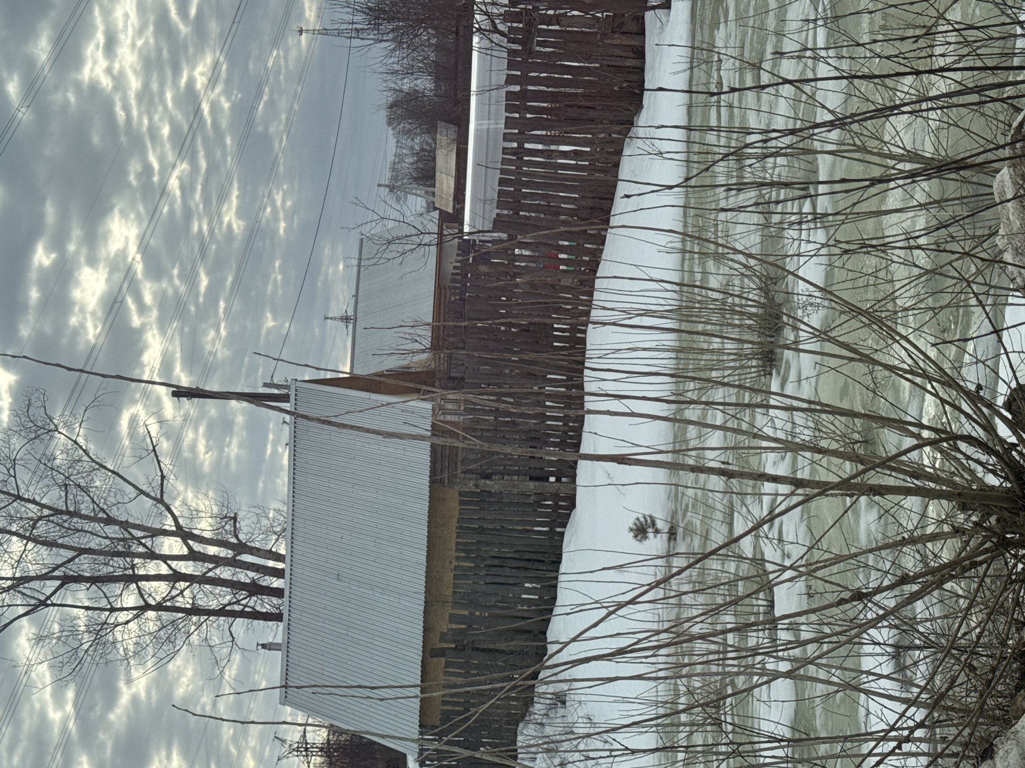 The image presents a scene in a likely rural area, under an overcast sky with textured clouds. A building with a corrugated metal roof stands to the left, adjacent to a wooden fence. Patches of snow cover the ground, and there is the suggestion of an icy or snowy body of water in the background. The foreground is filled with leafless branches. Electrical power lines are visible in the sky. The location is purported to be Chaikovsky, Russia. There are no people visible.