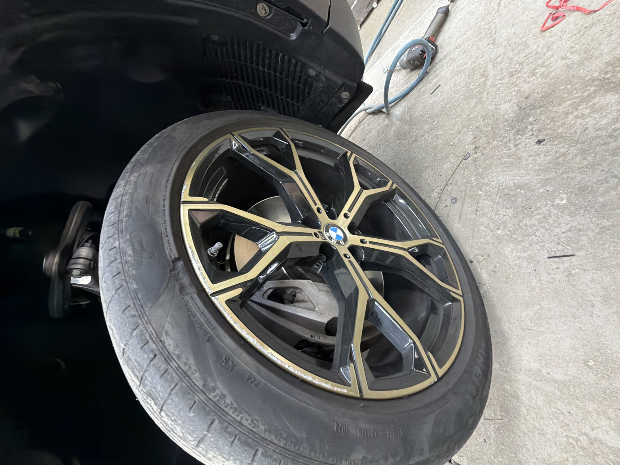 The image shows a close-up of a car's wheel and tire, which appears to be lifted off the ground, possibly for maintenance or repair. The wheel is black with gold accents and features a BMW logo in the center. The tire is partially visible and shows some wear. In the background, a car's suspension and fender are visible, along with a dark interior. On the ground, an air hose and a tool, possibly an impact wrench, are present, suggesting a workshop or garage setting. The floor is concrete and appears to be somewhat dirty, with some debris scattered around.
While the image captures a scene in what could be a garage, there is no visual information within the image to indicate its specific location in Burgas, Bulgaria. The context provided about Burgas is external to the visual data of the image itself.