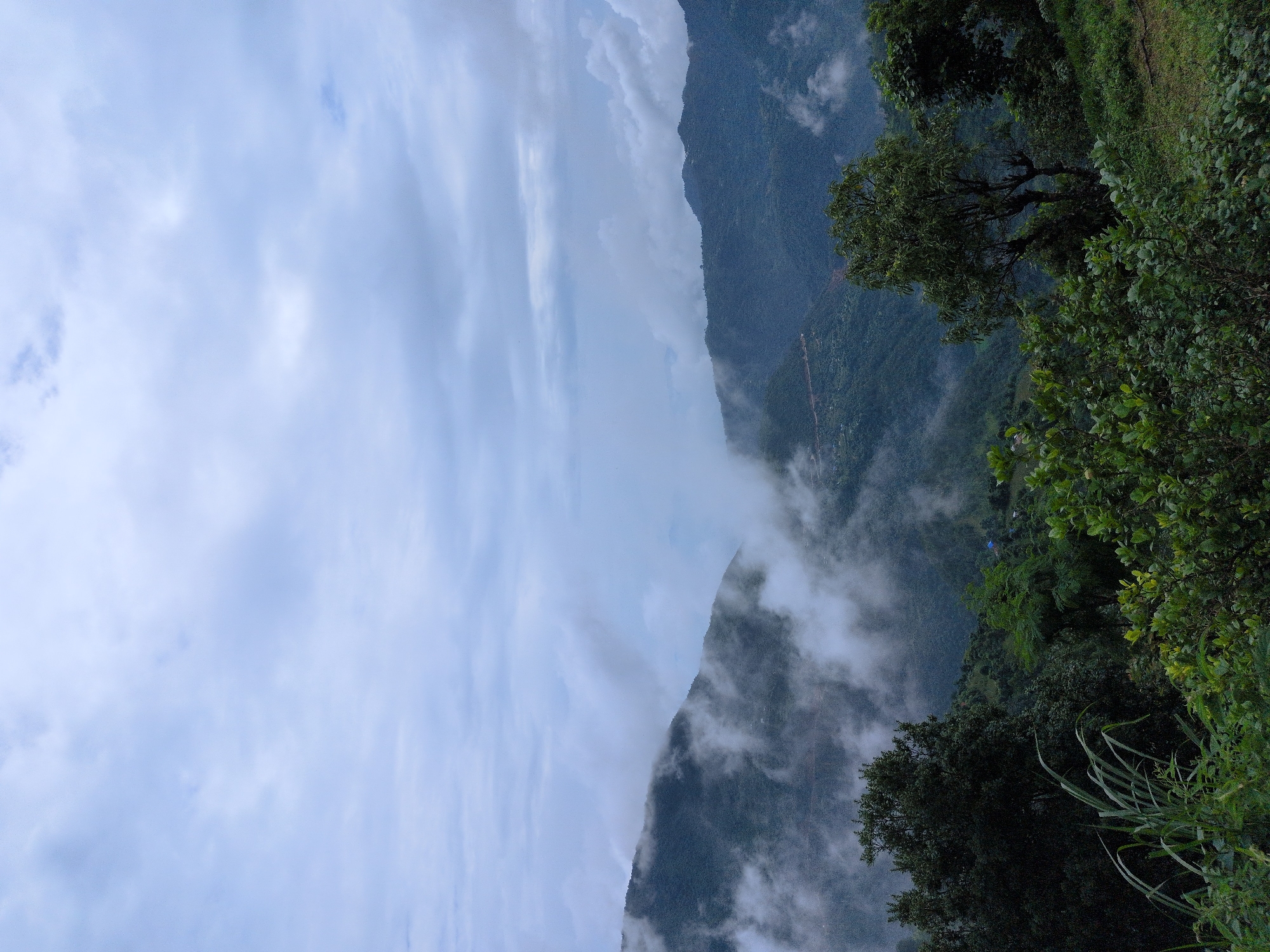 The image depicts a mountainous landscape on a cloudy day, likely in the region of Kapurkot, Nepal. Lush green vegetation covers the foreground, with various types of trees and bushes. In the mid-ground, rolling hills are partially obscured by mist and clouds, creating a serene and somewhat mysterious atmosphere. A winding road can be seen on one of the hillsides, suggesting human presence and connectivity in the remote area. The background features more distant mountains, also shrouded in clouds, which blend into the overcast sky. The overall scene is characterized by its natural beauty, with a focus on the interplay of land, vegetation, and atmospheric conditions. There are no people or animals visible in the frame.