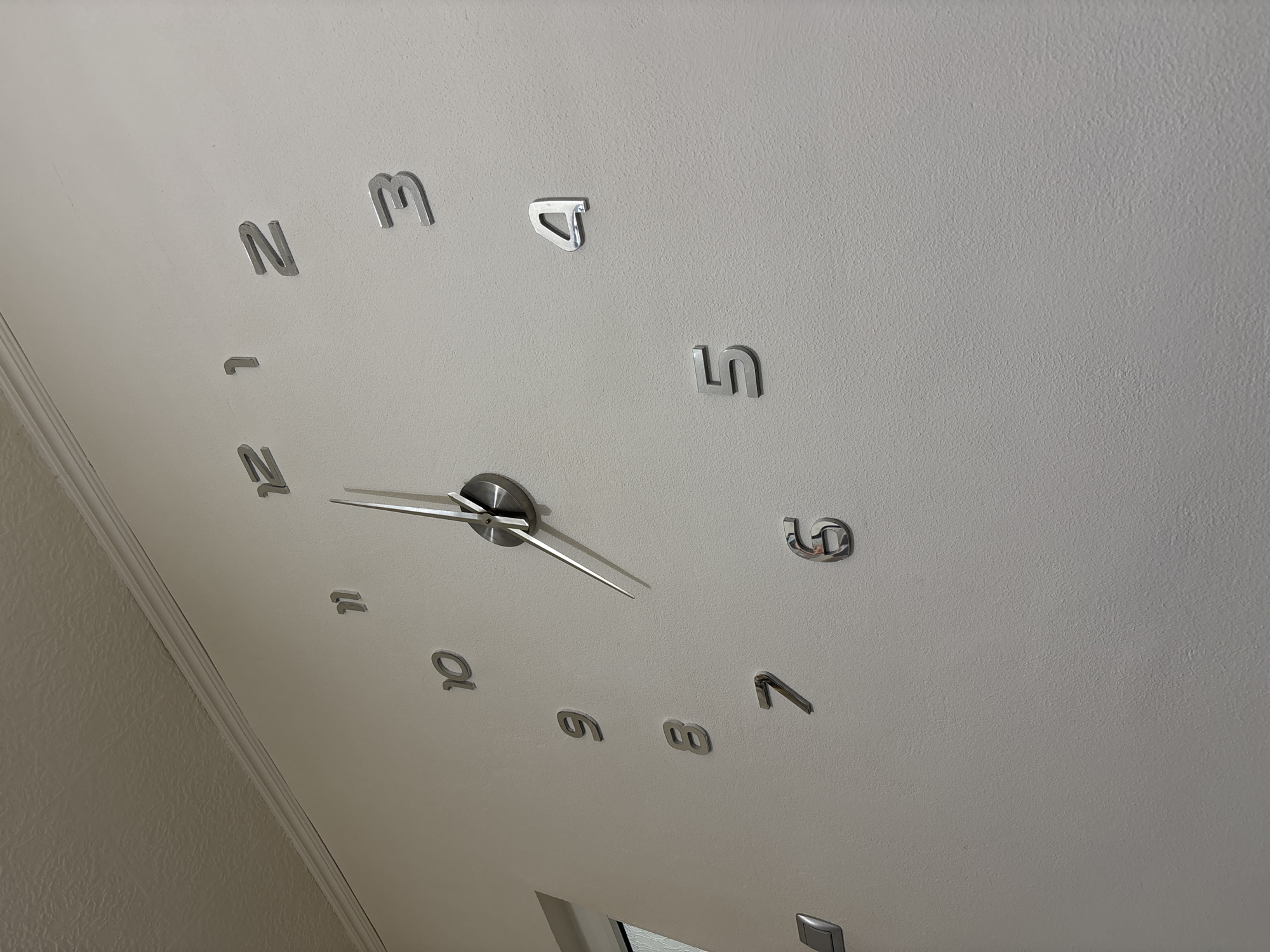 A low-angle, indoor shot focuses on a wall clock with large, reflective, sans-serif numeral markers. The numbers are arranged in a circular fashion, indicating the hours, with a reflective center mechanism holding the minute and hour hands. The minute hand points towards the number 5, while the hour hand is closer to the number 11. The environment appears to be a room with light-colored walls. It's not possible to locate the specific location in Plovdiv, Bulgaria, or describe any people or interactions based on this image alone.