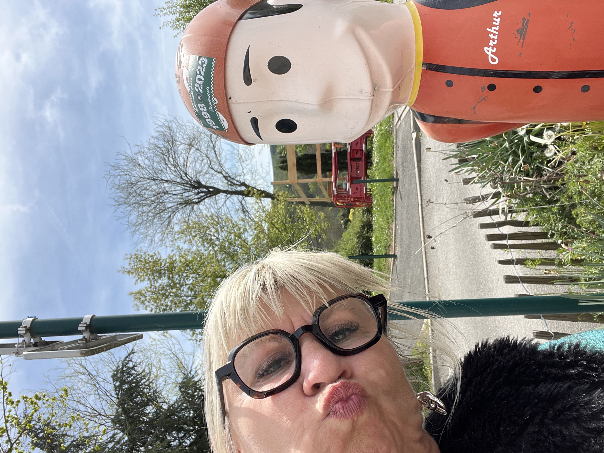 A playful selfie captures a woman posing with a large, cartoon-like statue outdoors. The subject in the foreground is a woman with short, light blonde hair and dark-rimmed, square-framed glasses, making a "kissy face" expression towards the camera. She appears to be wearing a dark, possibly faux fur, garment.
Above and slightly behind her is a prominent statue of a male figure. The statue has a light-colored, simplified face with black oval eyes and a straight line for a mouth. It wears an orange-red top with a yellow collar, black vertical stripes, and black circular details, and a matching orange-red helmet-like head covering.
The setting is an outdoor area, likely a park or a roadside in La Fouillouse, France, characterized by green trees and foliage under a bright, partly cloudy sky. A paved path or road is visible in the background, along with a wooden fence and some distant red structures.
Notable details include text visible on the statue: a green and white sticker on its helmet reads "1998 - 2023" and "Gargamels," while the name "Arthur" is written in white script on its orange-red torso. The image appears to be taken during daylight hours with good natural lighting, conveying a lighthearted and informal mood.