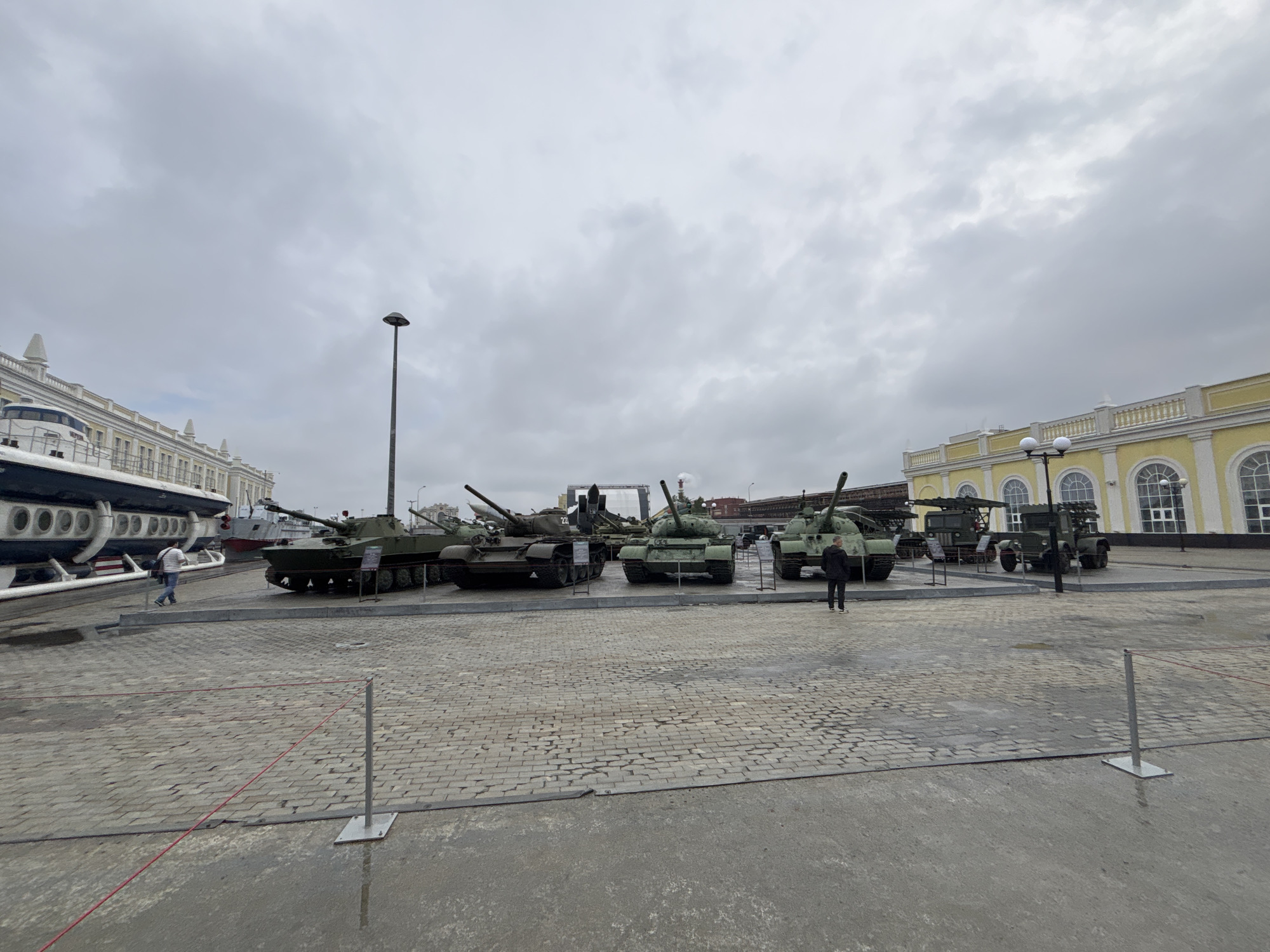 In Verkhnyaya Pyshma, Russia, an outdoor exhibit showcases military vehicles against a backdrop of overcast skies and a distinctive yellow and white building with arched windows. In the foreground, cobblestone paving is wet, reflecting the dull light, and a red rope barrier runs across the scene, supported by metal posts. To the left, a large, white, futuristic-looking vessel with a dark band runs along its length, is parked next to a display of several green military tanks. One tank is a smaller, lighter green variant, while the others are darker green. A person in a striped shirt and blue jeans walks away from the camera toward the tanks. In the middle ground, a series of larger, darker green military tanks are parked in a row, with some appearing to have additional equipment mounted on them. A man in a black jacket and pants stands to the right of these tanks, looking towards the vehicles. Further to the right, a green military truck with a rocket launcher system is parked. In the background, a factory with smokestacks is visible, along with more buildings.