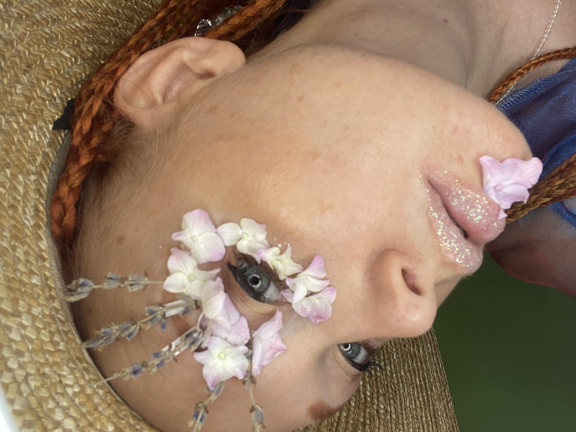 A close-up, low-angle shot focuses on a person's face, adorned with delicate flowers and glitter. The person is wearing a straw hat, and their hair is styled in braids. The background is a solid green color, suggesting an indoor or controlled outdoor setting. There is no indication of the city of Sochi, Russia, in the image.