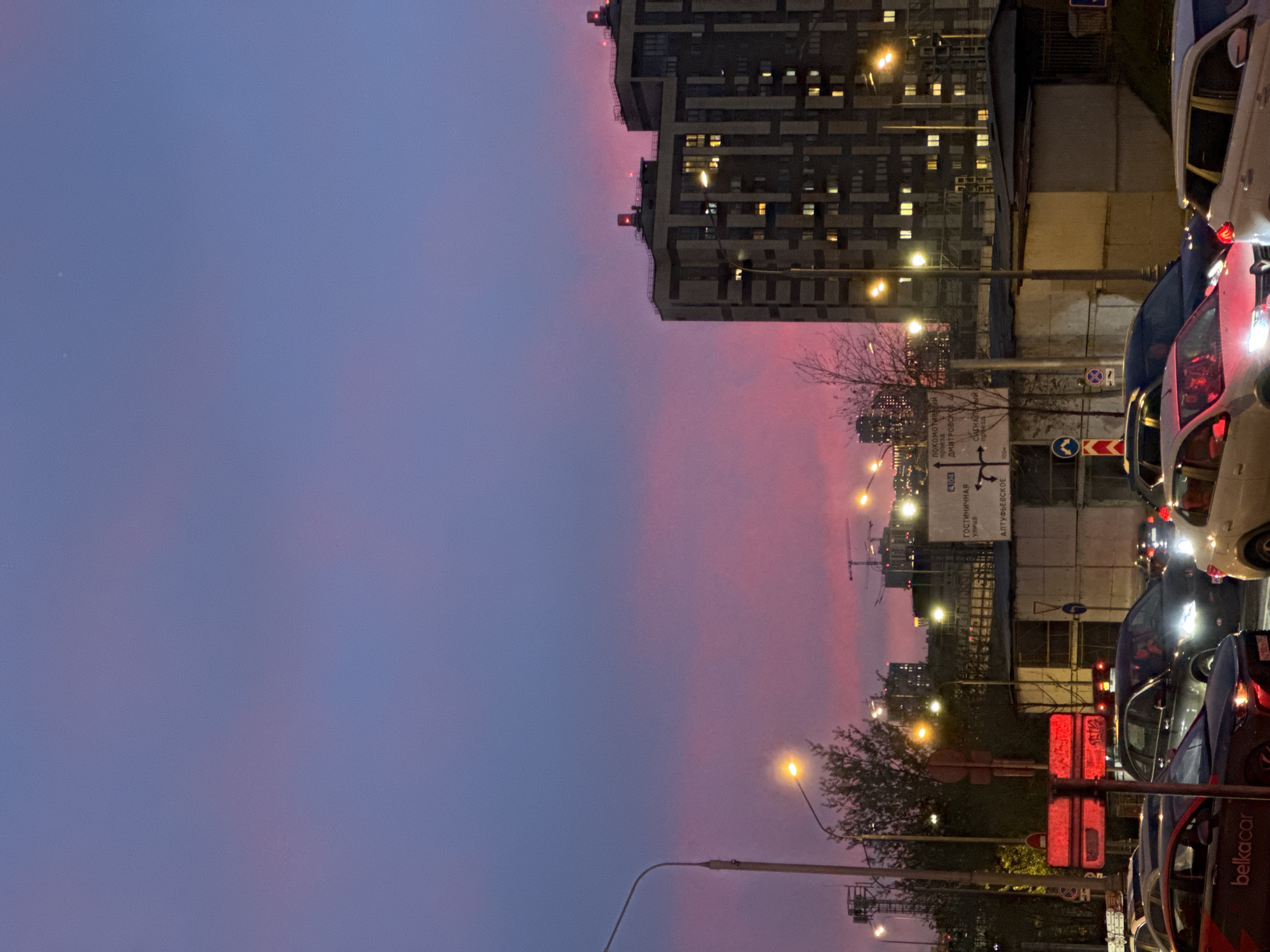 The image depicts a Moscow street scene at dusk. The sky transitions from a blue hue on the left to a pinkish-purple on the right. A tall, modern apartment building stands in the background on the right, with several illuminated windows. Streetlights are visible along the road. A traffic jam is present on the street, with several cars lined up. A sign with Russian text is also visible, pointing to different directions. The text "Алтуфьевское шоссе" (Altufyevskoye Shosse) and "Гостиничный проезд" (Gostinichny Proezd) can be read on the sign. A "belka car" sign is visible on a red car.