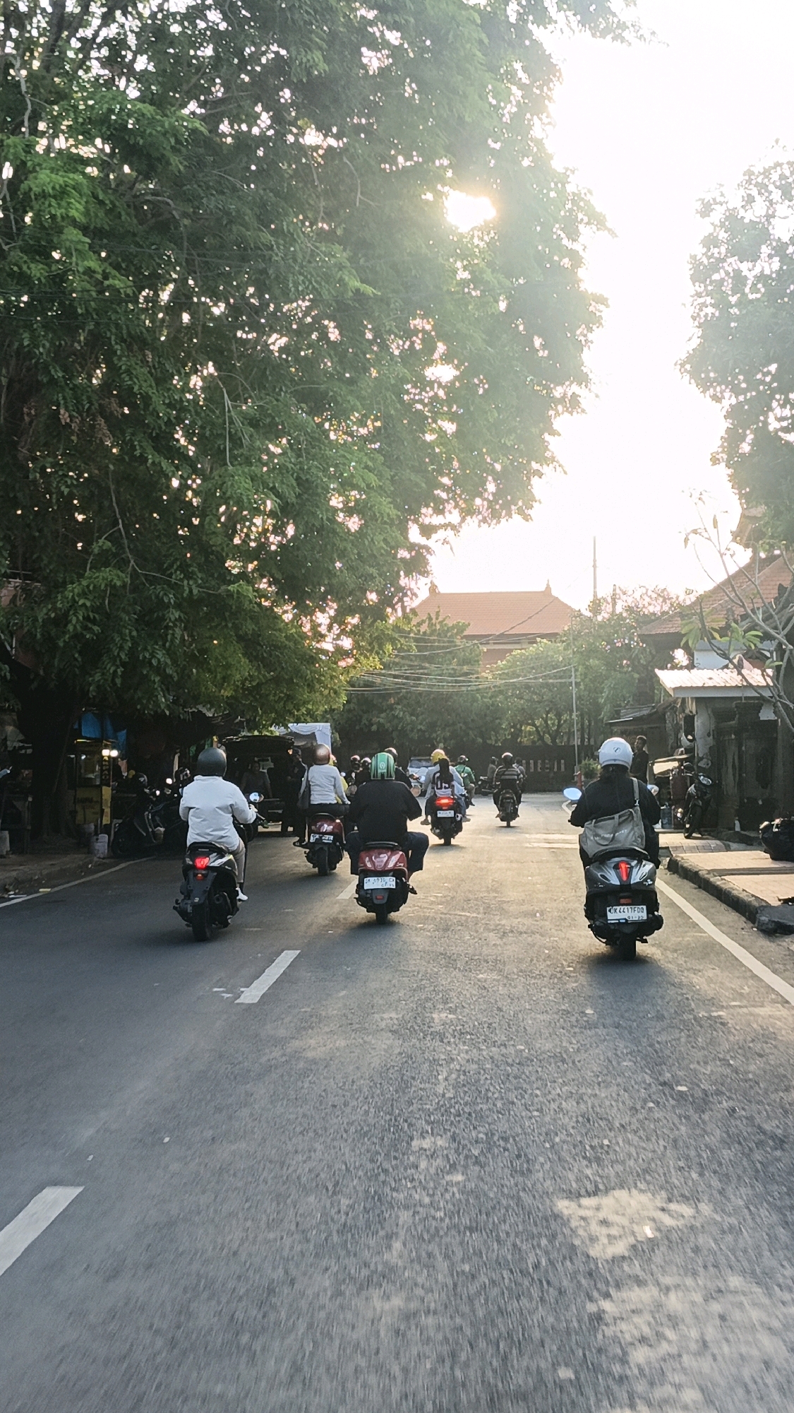 The image depicts a street scene in Denpasar, Indonesia, at dusk. The road is paved with asphalt and marked with white lines. A line of people on motorbikes is moving away from the viewer, heading towards a building with a traditional Balinese architectural style in the background. The sunlight filters through the lush green trees on the left side of the road, creating a dappled effect. On the right, there are shops and a sidewalk. The sky is bright and hazy, indicating the setting sun. The overall atmosphere is that of a typical evening commute in a tropical city.