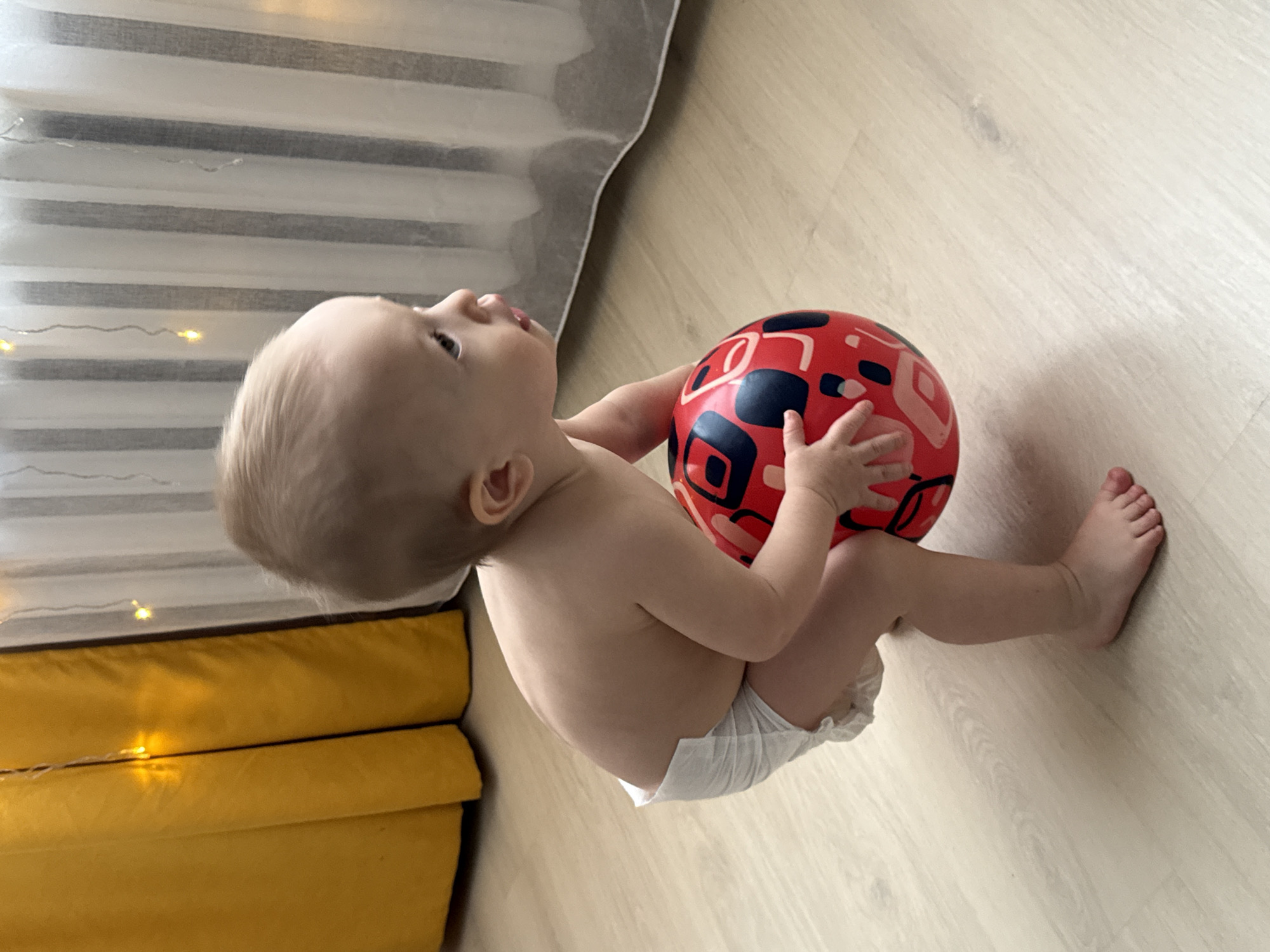 A baby wearing a diaper is sitting on a light-colored wooden floor. The baby is holding a red ball with black and red markings. Behind the baby is a yellow cushioned object and a white curtain with string lights. The location is possibly in the city of Chaikovsky, Russia.