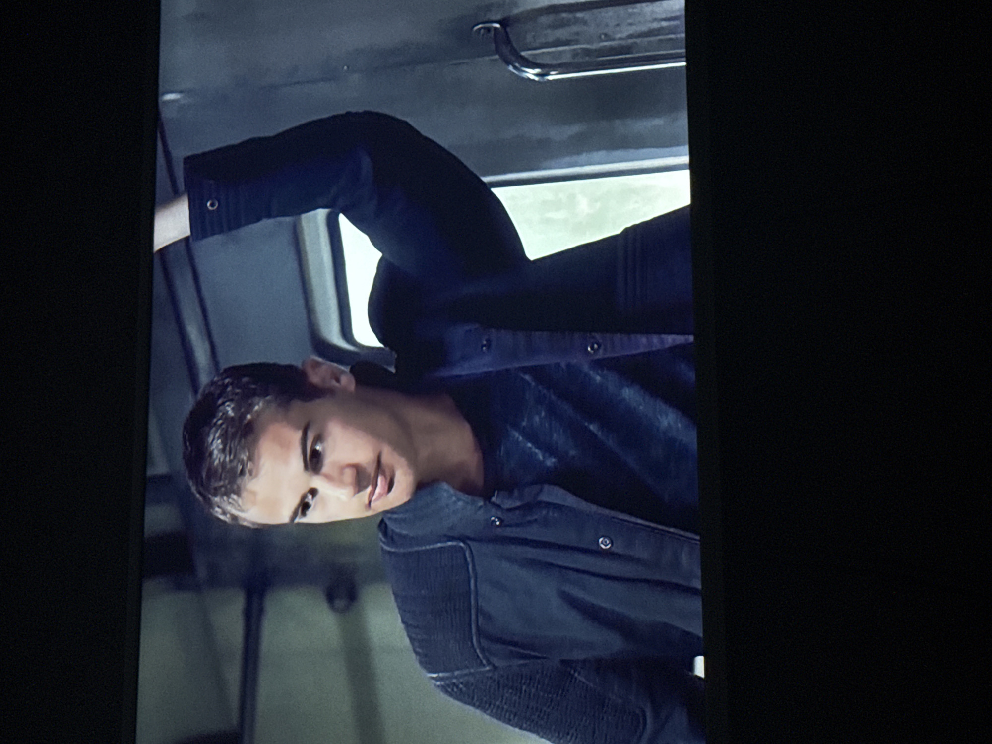 A young man stands inside what appears to be a train car or bus. He is wearing a dark blue jacket and a grey shirt. He is holding onto a metal handle above. The background shows a window with a view of greenery, suggesting the vehicle is in motion. The location cannot be determined from this image.