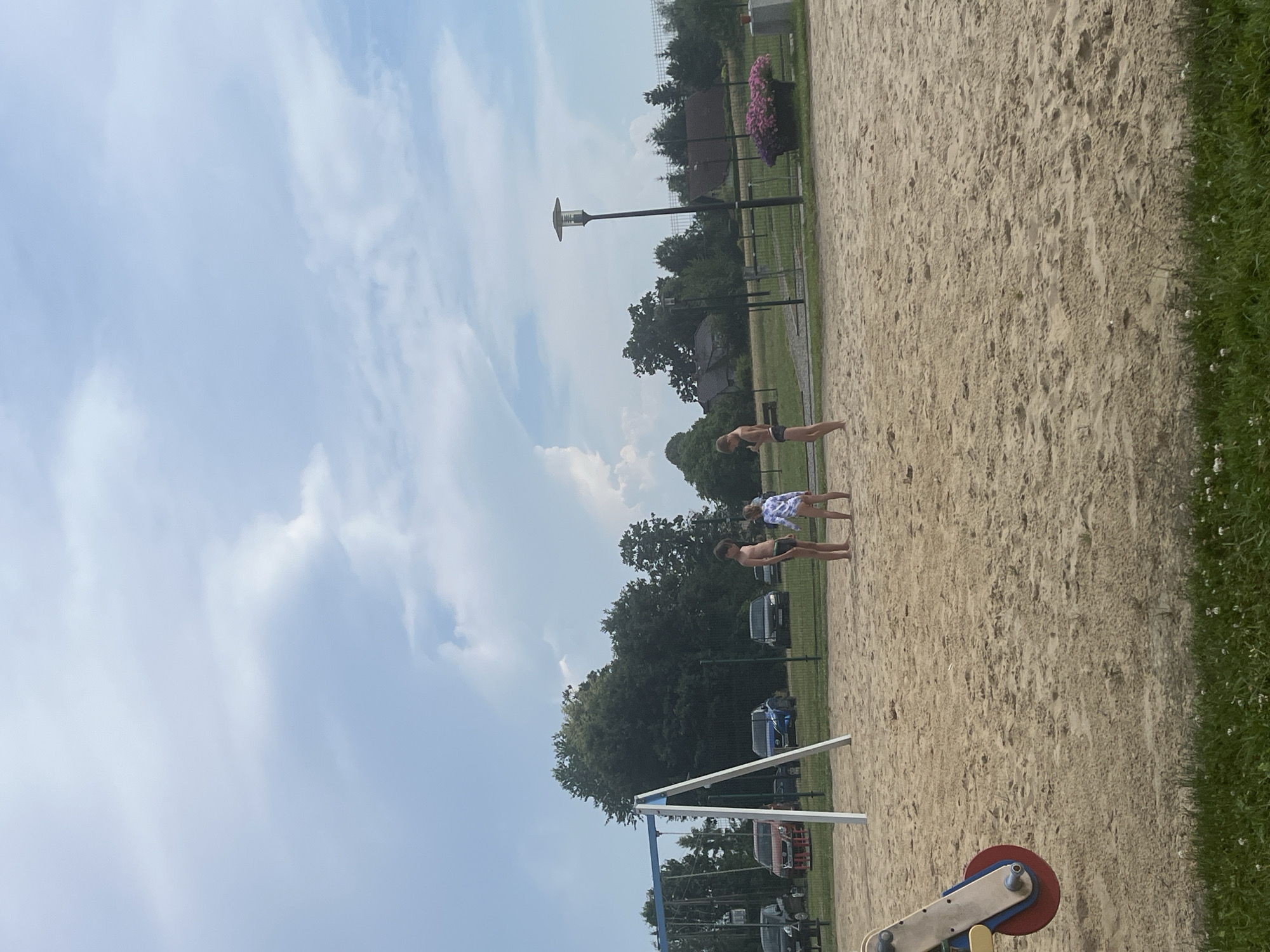 The image shows a sandy area, possibly a playground or beach, with three children standing and facing each other. A boy in black swimming trunks is on the left, a younger child in a patterned swimsuit is in the middle, and another boy in black swimming trunks is on the right. In the background, there is a swing set, several parked cars, trees, houses, a lamp post, and a large planter filled with pink flowers. The sky is partly cloudy.