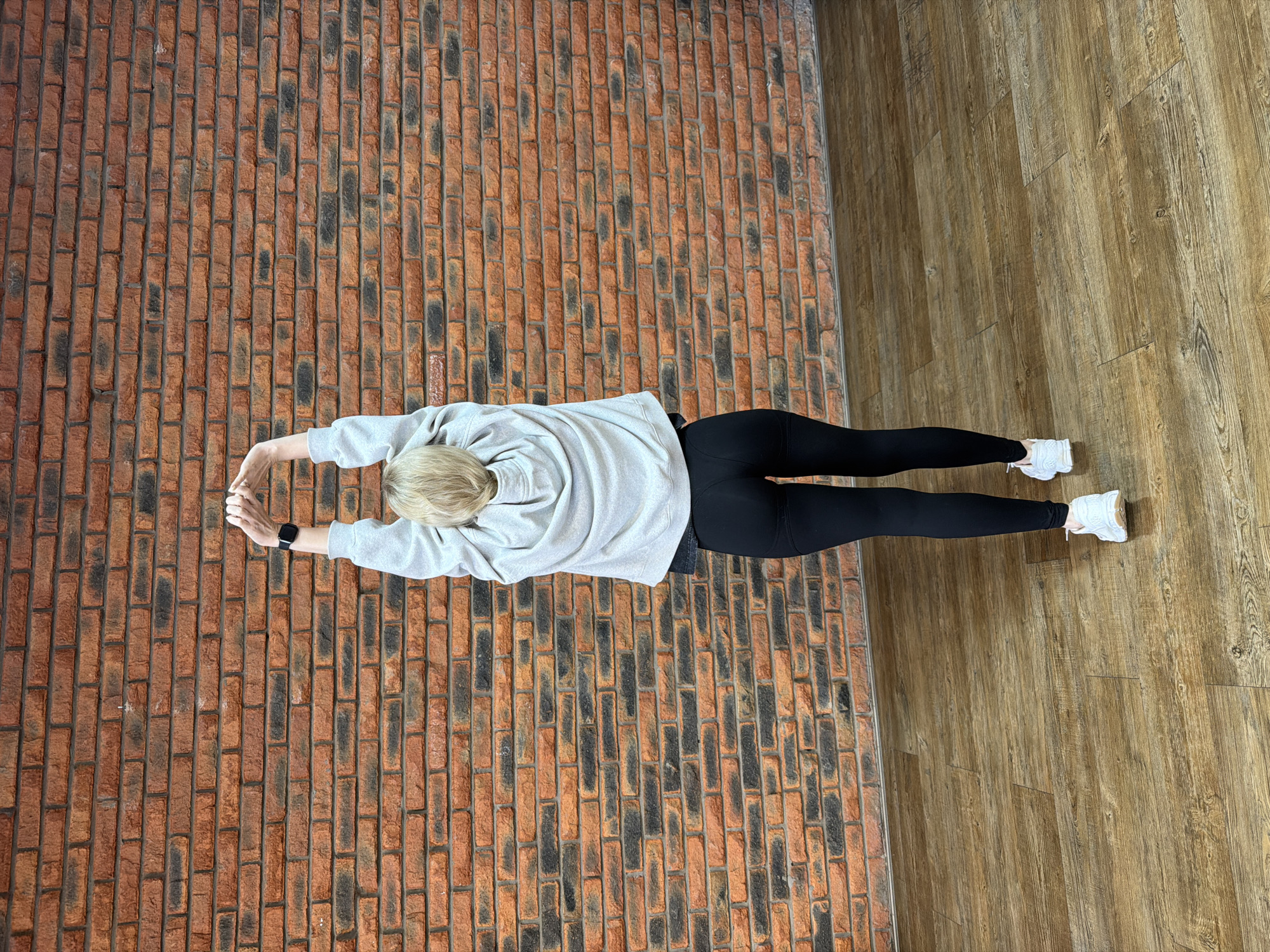 An overhead shot depicts a person lying prone on a split-surface floor. The individual, who has blonde hair and appears to be an adult, is dressed in a light gray hooded sweatshirt, black leggings, and white sneakers. A black smartwatch is visible on their left wrist. Their arms are extended forward and hands are clasped together, suggesting a stretching exercise or yoga pose.
The floor is distinctly divided into two sections. On the left side, there is a reddish-brown brick pattern, featuring dark grout lines and scattered darker, possibly aged or distressed, brick elements. On the right, the floor transitions to a light brown wood-look surface, laid in planks with visible grain details. The division between the brick and wood patterns forms a straight vertical line through the scene.
The setting is indoors, indicative of a gym, fitness studio, or a dedicated exercise area. The lighting is bright and evenly distributed, consistent with an interior environment. No specific time of day is discernible, and no text is visible within the image.