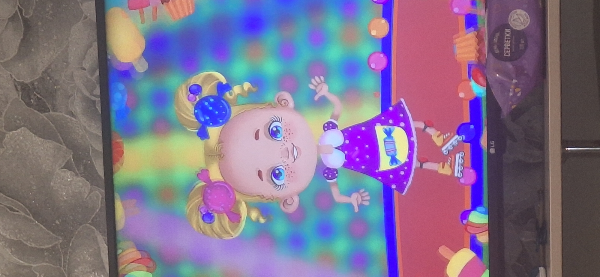 A stylized animated girl with blonde pigtails, blue eyes, and freckles stands on roller skates in front of a brightly colored, patterned background. She wears a purple polka-dot dress with a white collar and a yellow apron featuring a candy design. Her hair is adorned with candy-shaped hair clips, one pink and one blue. The background transitions through vibrant colors, featuring blurred circular shapes. To the lower right, a purple package with Cyrillic text, likely "СЕРБЕТКИ," meaning "wipes," is visible on a surface below the screen. The LG logo is present on the screen's lower bezel. The scene appears to be displayed on a television screen, with a textured wallpaper visible above it. The image does not provide any information about the city of Bila Tserkva, Ukraine.