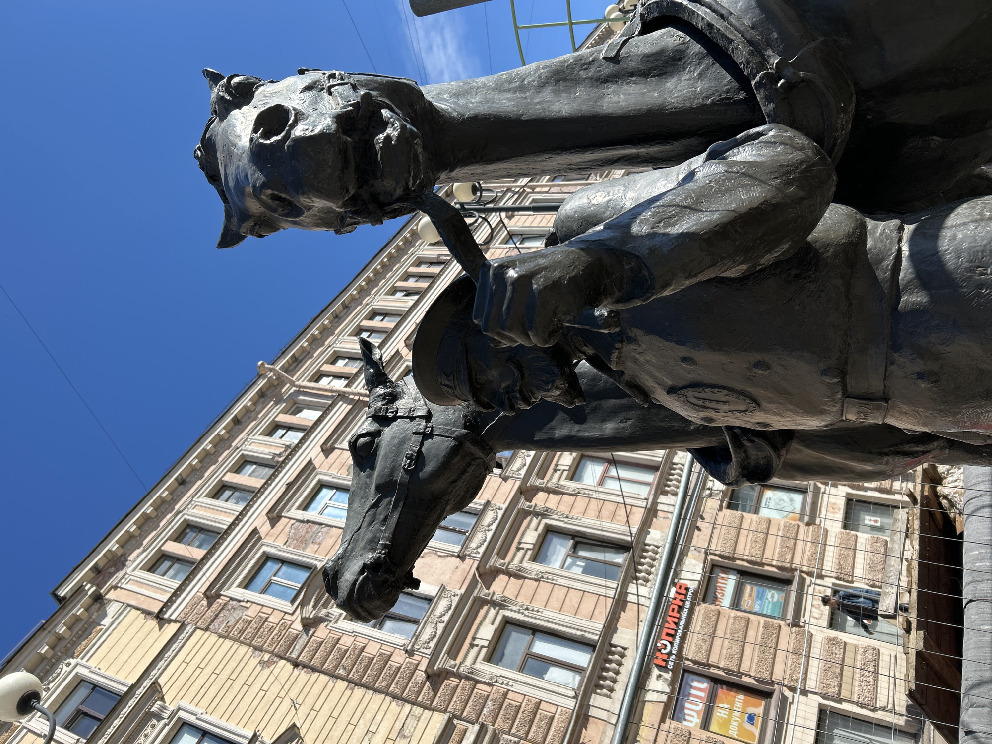 A low-angle, upward-facing shot captures a dark bronze equestrian statue against a clear blue sky, with a multi-story, ornate building forming the background. The statue depicts a person on horseback. The horse's head is prominently featured in the foreground, detailed with visible eyes, nostrils, and ears, and it wears a bridle. The rider, also rendered in dark bronze, is seen from the chest up, wearing a hat and what appears to be a uniform or formal attire, holding reins in their hands.
The building behind the statue is a classical or neoclassical structure, featuring a light-colored facade with intricate architectural details such as cornices, decorative pilasters, and textured wall sections. Numerous rectangular windows are visible across its many stories. The bright sunlight creates strong highlights on the statue's metallic surface and illuminates the building. The sky is a vibrant, cloudless blue, suggesting clear weather during the daytime. Thin wires, possibly for utilities or streetlights, stretch across the upper portion of the sky. A metal mesh fence or barrier is partially visible on the right side, close to the statue.
Text in Cyrillic is visible on banners on the building: "КОПИРКА" (Kopirka) and "СЕТЬ КОПИРОВАЛЬНЫХ ЦЕНТРОВ" (Network of Copy Centers), indicating a copying service. Another banner partially shows "Я ДОКУМЕНТ" (I am a document). The architecture and text cues confirm the location as Saint Petersburg, Russia.