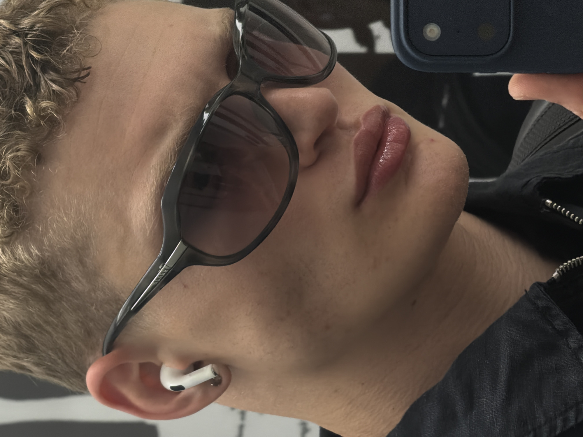 A close-up, profile shot captures a person wearing dark-rimmed sunglasses and a white wireless earbud in their left ear. Their short, blonde hair is styled with a textured, curly finish. The person is wearing a dark collared shirt, possibly a jacket, with a visible zipper. The background appears to be an indoor setting with a reflective surface, suggesting a mirror or polished wall, and hints of what could be a city environment, though no specific landmarks are identifiable. The lighting is somewhat diffuse, highlighting the textures of the skin and hair, and the sheen on the lips. The image is a selfie, as indicated by the partial view of a smartphone in the lower right corner. There are no discernible London landmarks or specific urban features to place this image within the city.