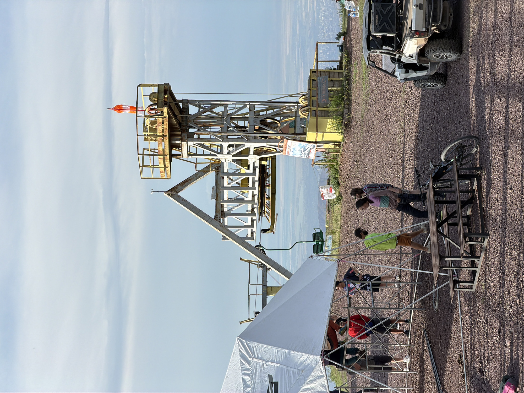 A ski lift station stands on a rocky hill with a panoramic view of a city in the distance, likely Kashka Suu, Kyrgyzstan. A white tent is set up to the left, with several people gathered around it. In the foreground, a picnic table and a bicycle are visible. A white all-terrain vehicle is parked to the right. The sky is partly cloudy, and the sun is casting shadows, indicating it is either morning or late afternoon. The ski lift is a prominent structure with a yellow platform at the top, topped with an orange and white windsock. The overall scene suggests a recreational area or event at a mountain resort.
