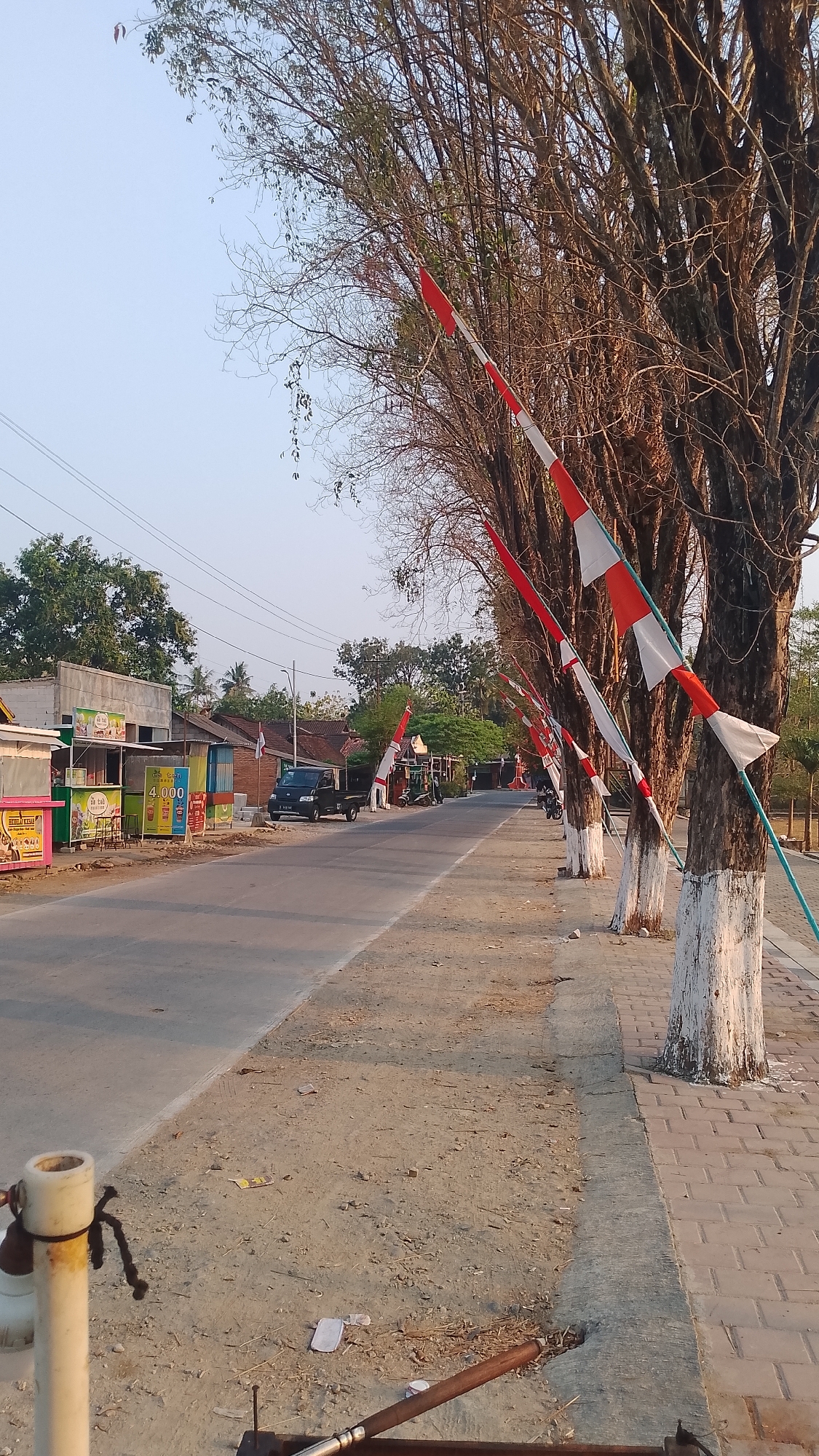 The image depicts a street scene in Karanglangu, Indonesia. The foreground features a dirt and gravel shoulder with some scattered litter, including what appears to be a white wrapper. To the left, a white pole with black cord attached is visible. The main subject of the image is a road lined with trees adorned with red and white Indonesian flags, tied to poles that are painted white at their bases. The trees are bare, suggesting a dry season. On the left side of the road, there are several small businesses or stalls with colorful awnings and signage. One stall has a sign with "4.000" on it. A dark-colored pickup truck is parked on the side of the road. Further down the road, more buildings and trees can be seen, with more flags lining the street. The sky is clear and light, indicating it is likely daytime. The overall impression is a quiet, semi-rural roadside with festive decorations.