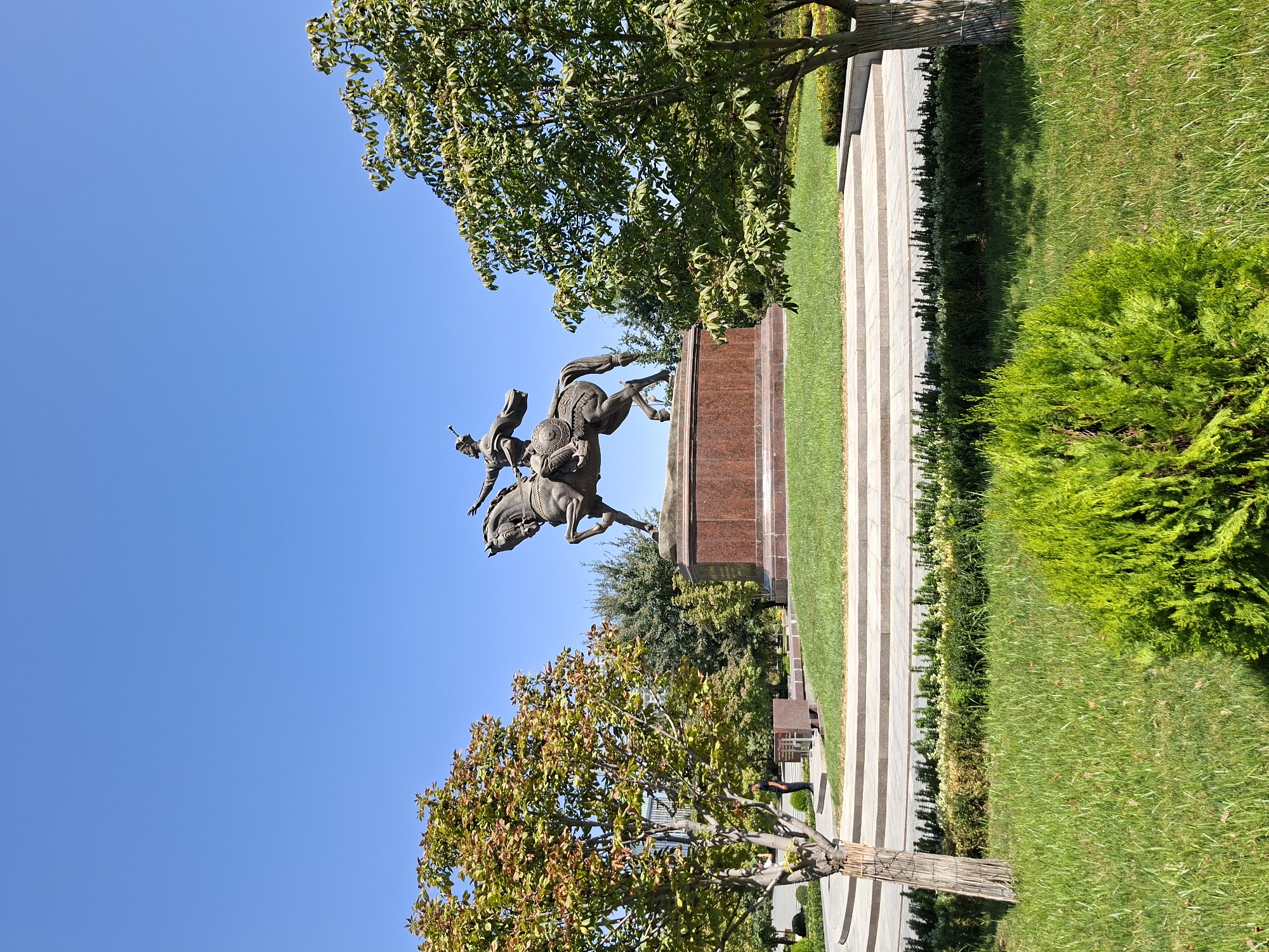 A statue of a man on horseback stands prominently in a park on a clear, sunny day. The statue is made of dark metal and depicts the rider in traditional attire, with a sword at his side and his arm outstretched. The horse is rearing on its hind legs. The statue is mounted on a tall, reddish-brown pedestal. Surrounding the pedestal is a circular lawn with meticulously maintained green grass. Behind the statue, a cluster of trees creates a backdrop against the bright blue sky.
In the foreground, the park features lush green grass and manicured bushes. To the left and right of the statue, two trees with a mix of green and reddish-brown leaves provide framing. A set of wide, light-colored stone steps leads up to the elevated platform where the statue resides. A person in dark clothing stands near a low wall and bench to the left of the steps. The ground is paved with stone in the lower portion of the image, with a neatly trimmed hedge line visible. The overall environment suggests a public park or square.