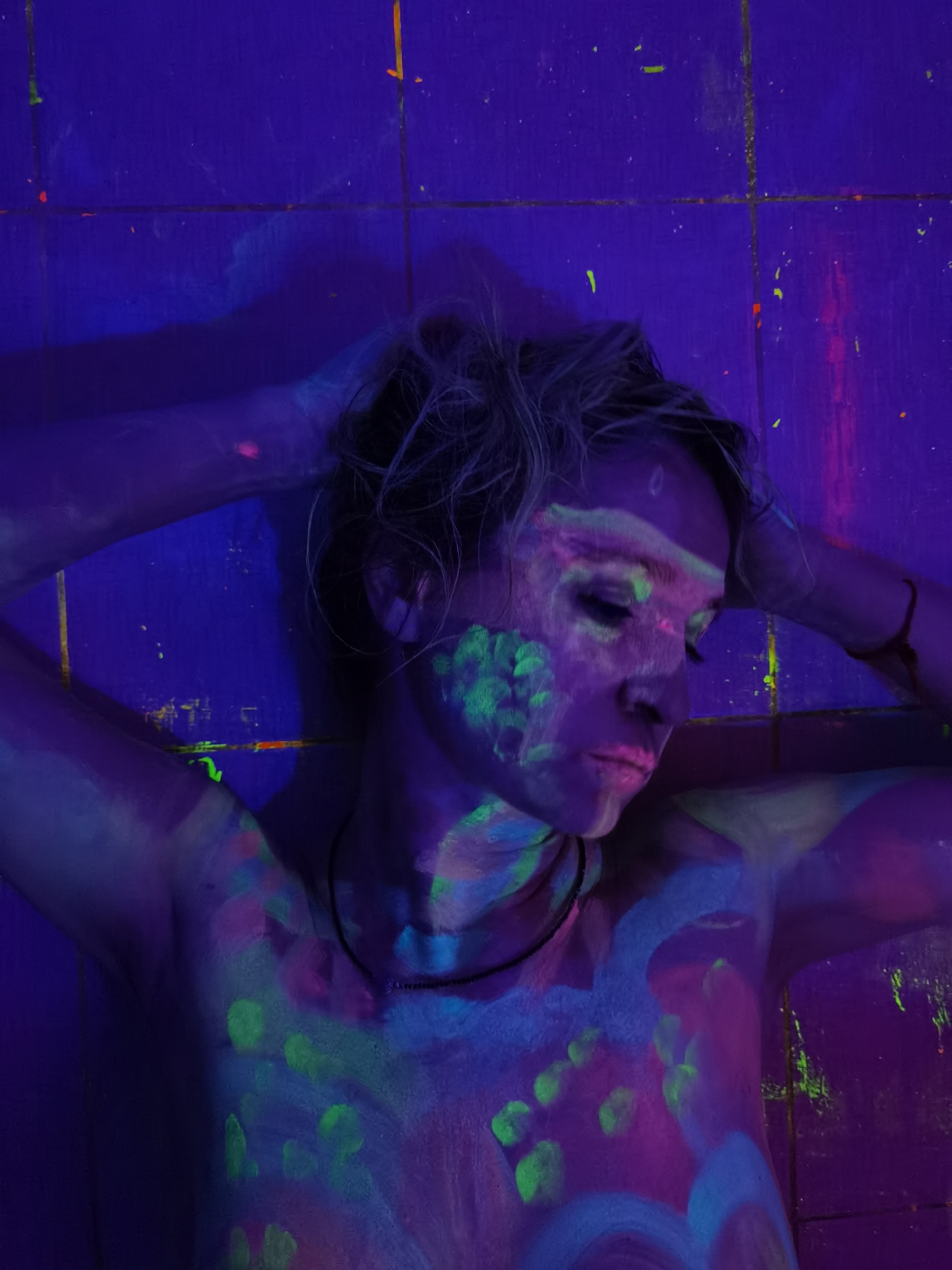 A person is depicted in a dark environment illuminated by UV (black) light, causing fluorescent body paint to glow. The subject, likely female, is shown from the chest up, lying down with her head turned slightly to the right, and her eyes closed or looking downwards. Her arms are raised, with hands resting near her head. Her light-colored hair appears bluish-purple under the UV light.
Her face, neck, and upper torso are covered in a vibrant array of glowing body paint in neon green, pink, and light blue. The patterns are abstract, featuring splatters, swirls, and what appear to be multiple glowing fingerprints, particularly on her chest and side. A thin, dark necklace is visible around her neck.
The background consists of a dark blue/purple tiled surface, possibly a floor or wall, with faint grout lines visible. There are also small splatters and streaks of the glowing paint on this surface. The overall atmosphere is artistic and ethereal, emphasizing the glowing body art. There is no visible text.