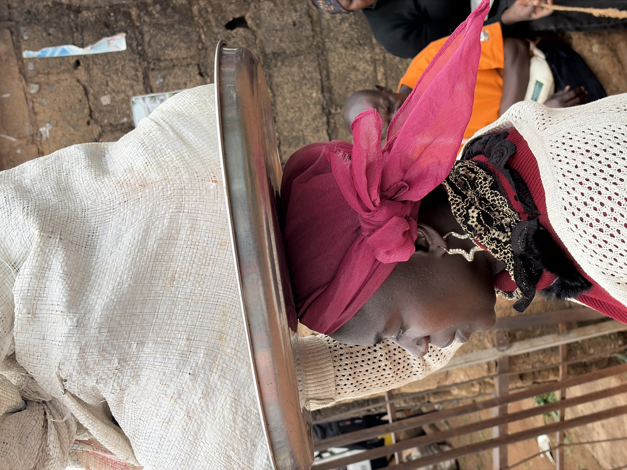 This is a close-up, vertically oriented image of a person's head and upper body. The subject is wearing a deep pink headscarf tied at the top of their head, and a cream-colored, loosely woven sweater or shawl over their shoulders. Beneath the shawl, other layers of clothing are visible, including what appears to be a dark red garment and black fabric with gold embellishments. A large, metallic tray runs vertically along the left side of the frame, partially obscuring the subject's body. The background is a mix of rough, textured ground and a metal railing.
The person's face is in profile, turned downwards. They have dark skin and are wearing large, ornate hoop earrings. The expression is neutral, with eyes closed or looking down. The lighting suggests it might be daytime, possibly outdoors, given the rough ground texture. The overall impression is of a candid, intimate portrait focused on the subject's attire and facial features. The image appears to have been taken in Gura Top, Nigeria, though no specific landmarks confirm this.