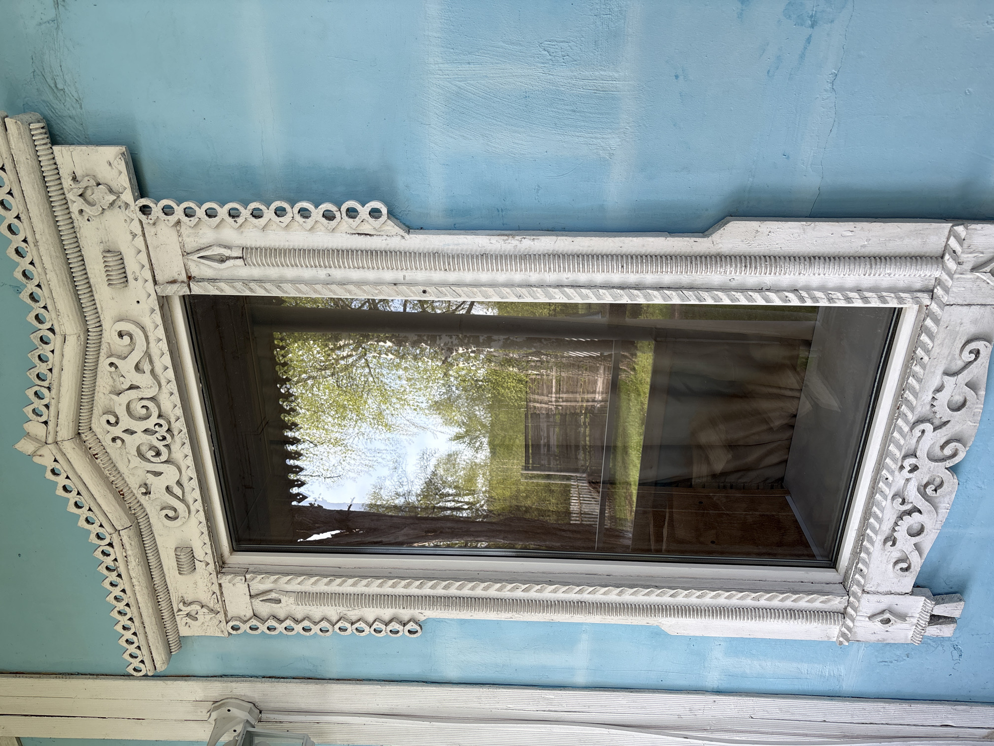 A close-up shot oriented on its side depicts an ornate window frame on a light blue exterior wall, potentially in Zavod Mikhailovskii, Russia. The window frame is painted white and features intricate carved details including scrolled patterns, scalloped edges, and linear indentations. The window pane itself reflects trees and a building, providing a glimpse of the outside environment. A horizontal white trim is visible at the bottom of the frame, indicating additional architectural elements.