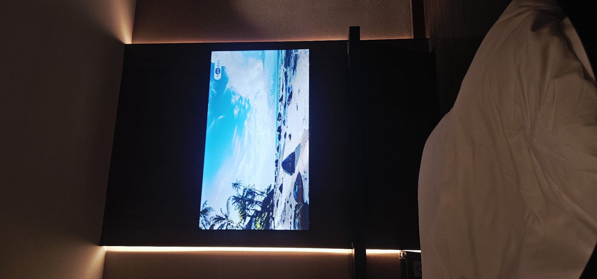 A partially lit television screen displays a beach scene with palm trees and blue water. The screen is mounted on a dark wall with an illuminated strip at the bottom. To the right, a bed with white bedding is partially visible. The location is possibly in Liangjiang, China, as requested, but there is no explicit information to confirm this.