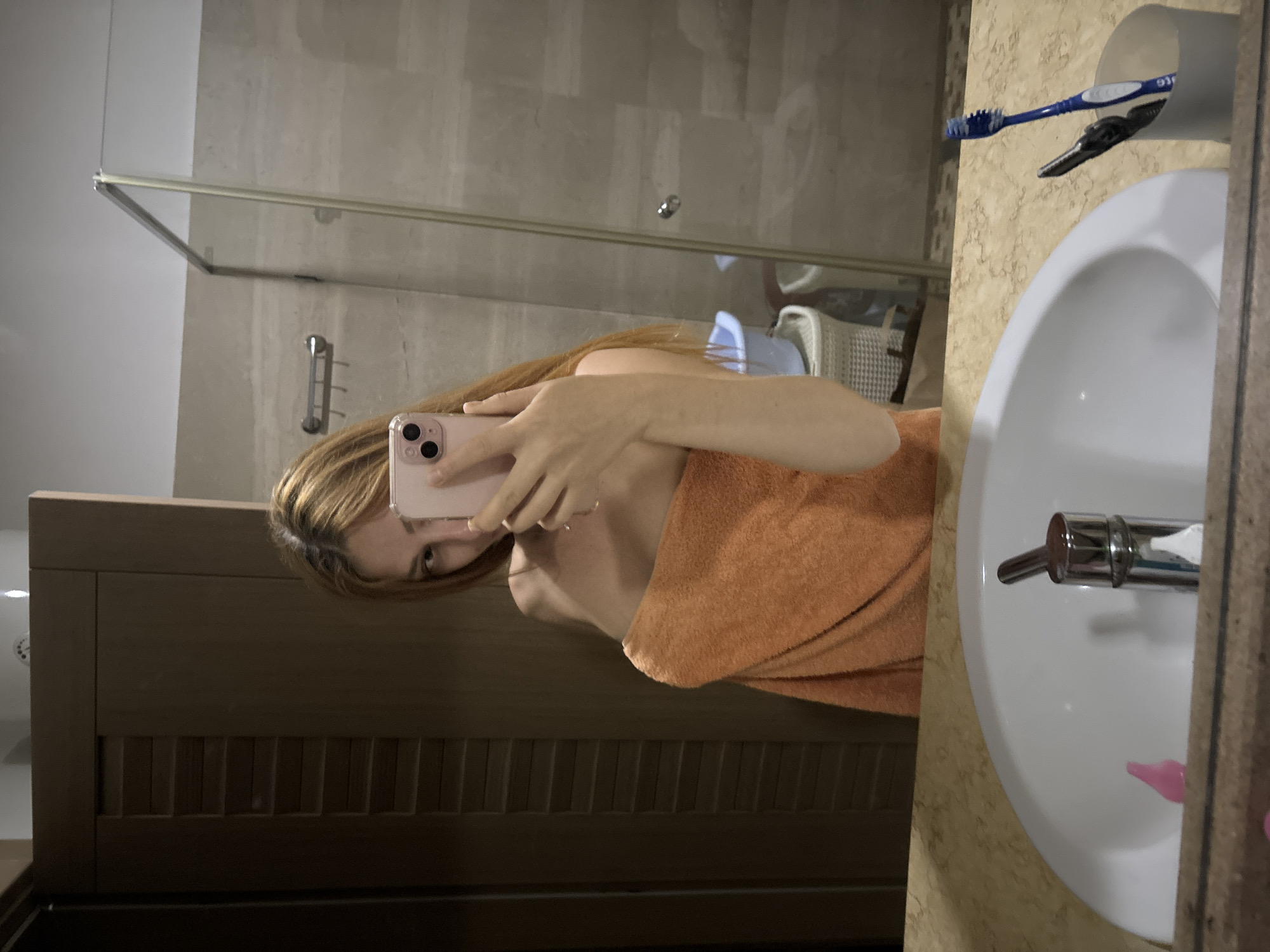In Budva, Montenegro, a woman is taking a mirror selfie in a bathroom. She is wrapped in an orange towel, and her hair is blonde. She is holding a pink smartphone. The bathroom has a sink, a faucet, a mirror, and a shower enclosure. The walls are tiled, and there is a wooden door in the background. There are also two toothbrushes and a pair of tweezers in a cup on the counter.