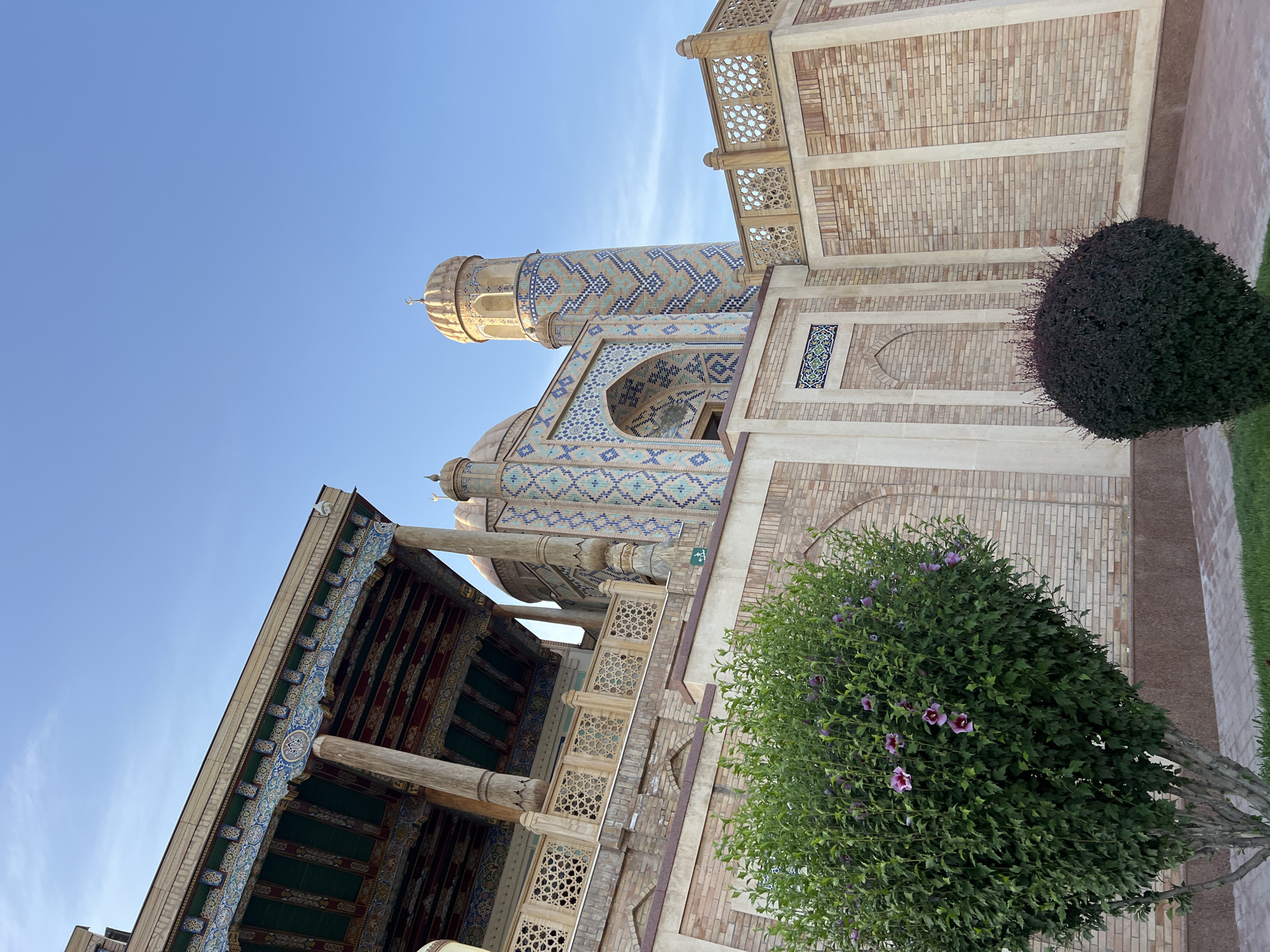 This is a low-angle, full exterior shot of the Chor Minor Madrasah in Samarkand, Uzbekistan. The madrasah, built in 1807, features four minarets topped with blue domes. The building's facade is decorated with intricate turquoise and blue mosaic tiles, and it sits on a raised platform made of light-colored brick. A large green bush with purple flowers is in the foreground to the left, and a smaller, dark green, spherical bush is to the right. The sky is clear and blue.