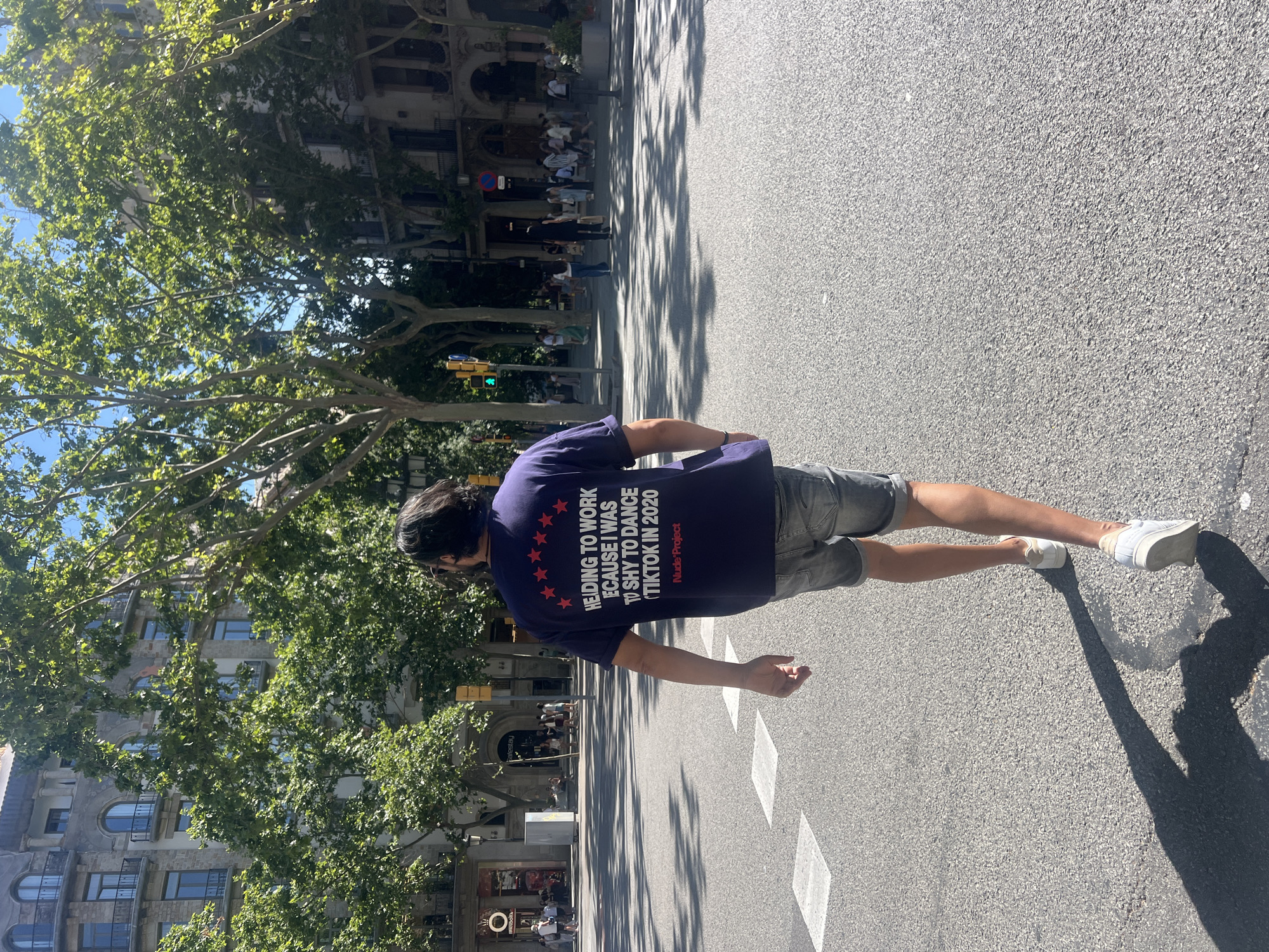 A man with dark, wavy hair walks away from the camera on a city street in Barcelona, Spain. He is wearing a dark blue t-shirt with white text and a graphic of red stars above it. The text on the shirt reads, "HEADING TO WORK BECAUSE I WAS TOO SHY TO DANCE (TIKTOK IN 2020)" and below that, "Nude Project". He is also wearing light wash denim shorts and white sneakers. The street is paved with asphalt and has white pedestrian crossing markings. In the background, there are buildings with traditional architecture, trees with lush green foliage, and other pedestrians. The sky is clear and blue. The scene is lit by bright sunlight, casting long shadows across the street.