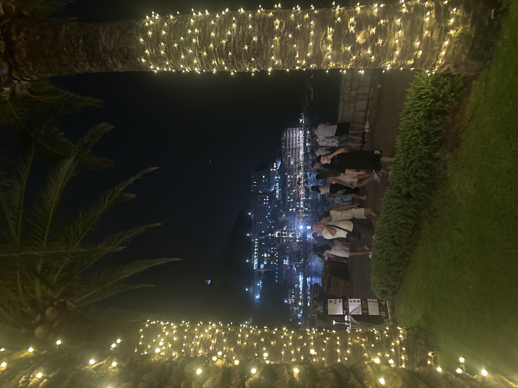 The image depicts a nighttime scene on a promenade in Bečići, Montenegro, illuminated by strings of warm-toned lights wrapped around palm trees. In the foreground, two large palm trees flank the view, their trunks adorned with a dense array of fairy lights. A grassy area and a neatly trimmed hedge are visible between the palm trees and a stone wall. A group of people, dressed casually, are walking and standing along the promenade, some interacting with each other. Beyond them, the coastal town of Bečići is visible, with buildings climbing up a hillside, their windows emitting a warm glow. The sea is present in the middle ground, with a few boats dotted on its surface. The overall atmosphere is festive and tranquil.