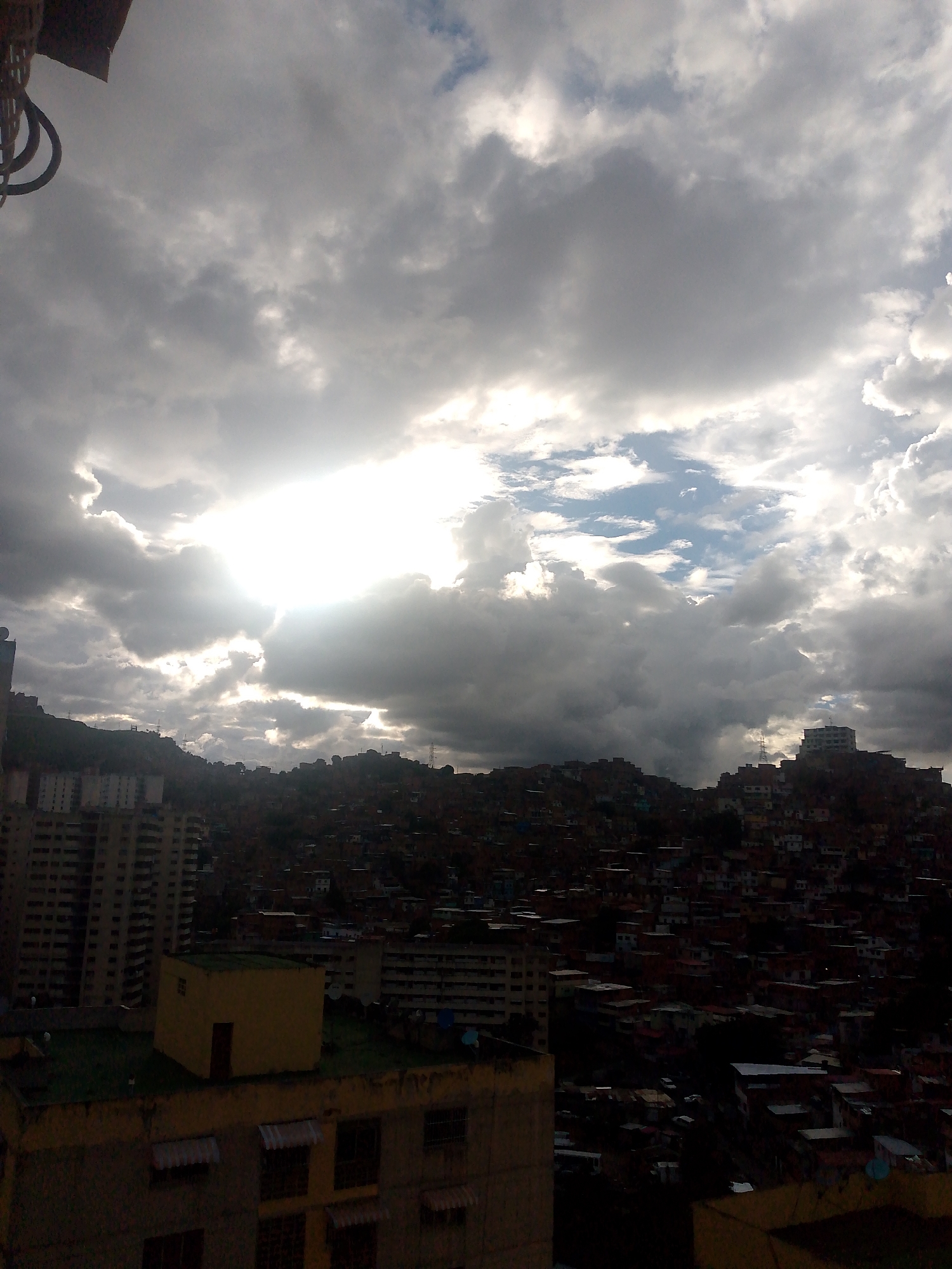 The image shows a view of a densely populated hillside neighborhood in Caracas, Venezuela, under a dramatic sky filled with dark, heavy clouds and patches of bright sunlight breaking through. The hillside is covered with numerous small, densely packed houses, many with red-tiled roofs, creating a mosaic of colors against the green of the vegetation and the darker hues of the buildings. In the foreground and midground, there are several multi-story buildings, some appearing to be apartment complexes with visible windows and balconies, while others are smaller, more utilitarian structures. One building in the foreground is yellow with a green rooftop. The overall impression is one of a sprawling urban landscape clinging to a steep incline, with the dynamic sky casting an intense light and shadow play across the scene. There are no people visible in the image. The photograph appears to have been taken from a higher vantage point, looking down and across the city.