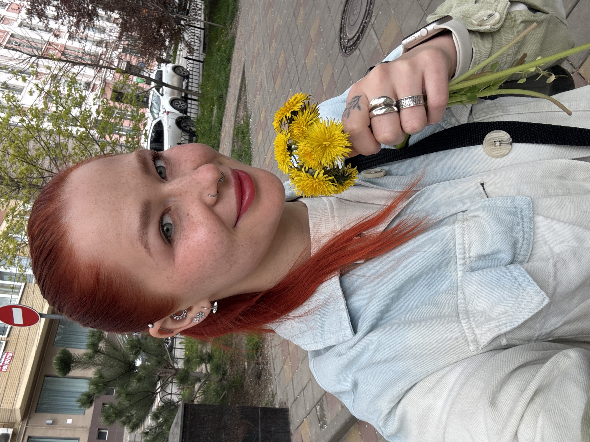 A young woman with red hair and multiple ear and nose piercings is holding a bunch of dandelions in her hand. She has freckles and is wearing several rings on her fingers. She is wearing a light blue denim jacket. The background shows a city street with trees, buildings, parked cars, and a sidewalk. A red and white road sign is visible. The location is suggested to be Rostov-on-Don, Russia, based on user-provided context.