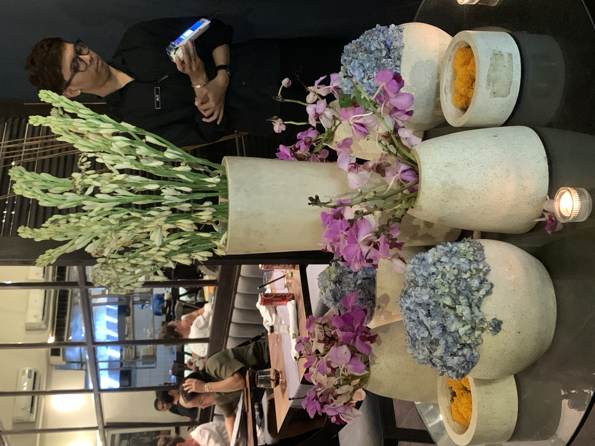 Captured at a restaurant in North Kuta, Indonesia, the image portrays an interior scene dominated by a table laden with floral arrangements and a person in a dark uniform.
The floral arrangements are the central focus. They comprise white, tall tubular flowers and a mix of purple orchids and blue hydrangeas, displayed in a variety of white, stone-like planters of differing shapes. One planter holds a vibrant yellow floral arrangement.
The top portion of the frame features a person in a dark uniform, wearing glasses and holding what appears to be a digital device. The person's gaze is directed towards the device.
The reflection in the tabletop reveals the restaurant's interior, showcasing additional patrons seated at tables. A Coca-Cola box and glasses are visible on one of the tables. Air conditioning units are installed on the walls. The lighting appears dim, suggesting an evening setting.