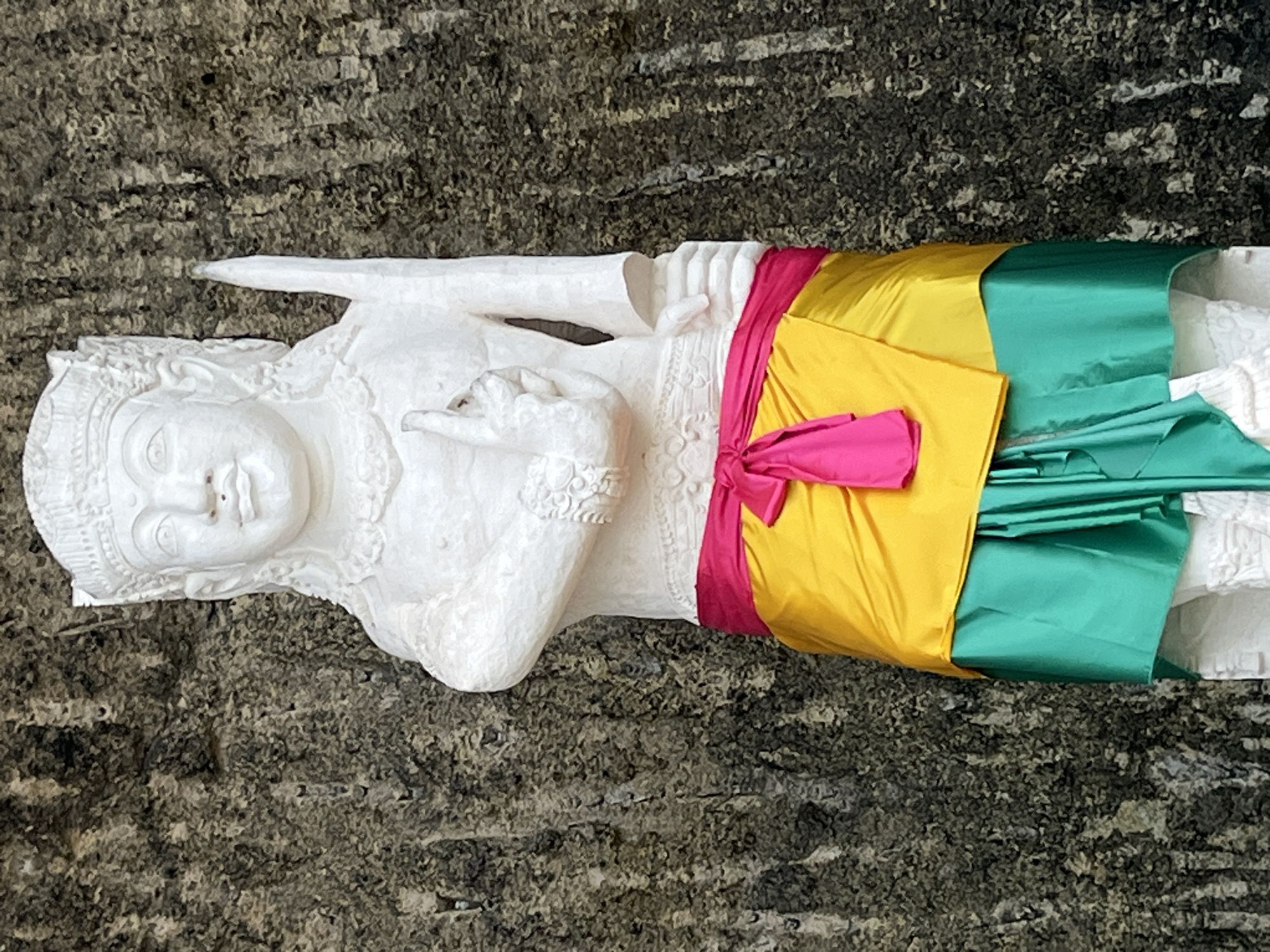 A white stone statue of a figure stands against a rough, textured, grey rock face in Kuta Selatan, Indonesia. The statue is adorned with colorful fabric. A bright pink sash is tied around the waist, over a flowing yellow sarong. Beneath the yellow sarong, a teal green skirt is visible, with pleats and folds. The figure's face is serene, with carved features and a headdress. The left arm is bent, with the hand raised and fingers pointing upwards in a gesture. The right arm is positioned downwards, holding a slender, pointed object that resembles a sword or dagger. The overall scene suggests a cultural or religious artifact in an outdoor setting.