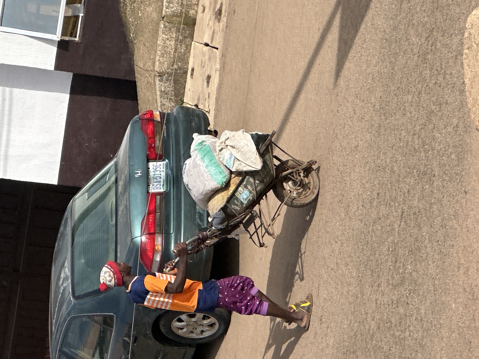 A young male, wearing an orange and blue striped t-shirt, purple patterned trousers, and a red and white patterned beanie, pushes a loaded wheelbarrow down a dirt road. The wheelbarrow is filled with sacks of what appear to be goods. Behind him, a dark green sedan is parked. The vehicle has a license plate that reads "BSA-5135C". The background shows a building with white and dark brown walls and a window. The scene is illuminated by sunlight, casting a shadow of the wheelbarrow and the young male on the ground. The image was taken in Bukuru, Nigeria.