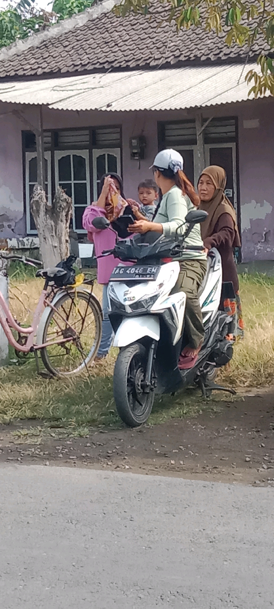 A group of four people are gathered outdoors in front of a building, near a paved road. The building appears to be a residential house with a tiled roof and a light purple exterior. A white motorcycle with a license plate reading "LAC 4046 EH" is parked in the foreground, with one person sitting on it. A pink bicycle with a basket is to the left of the motorcycle.
On the motorcycle, a person wearing a light green long-sleeved shirt and olive-green pants is seated. A child, possibly a toddler, is being held by another person to the left of the motorcycle. This person is wearing a bright pink long-sleeved shirt and has their hands covering their face, suggesting distress or crying. Another adult, wearing a brown hijab and a patterned skirt, stands to the right of the motorcycle, looking towards the person in pink.
The ground is covered in dry grass and scattered stones. The environment suggests a rural or suburban setting. The overall scene depicts a moment of interaction or concern among the individuals, with the presence of two vehicles indicating potential travel.