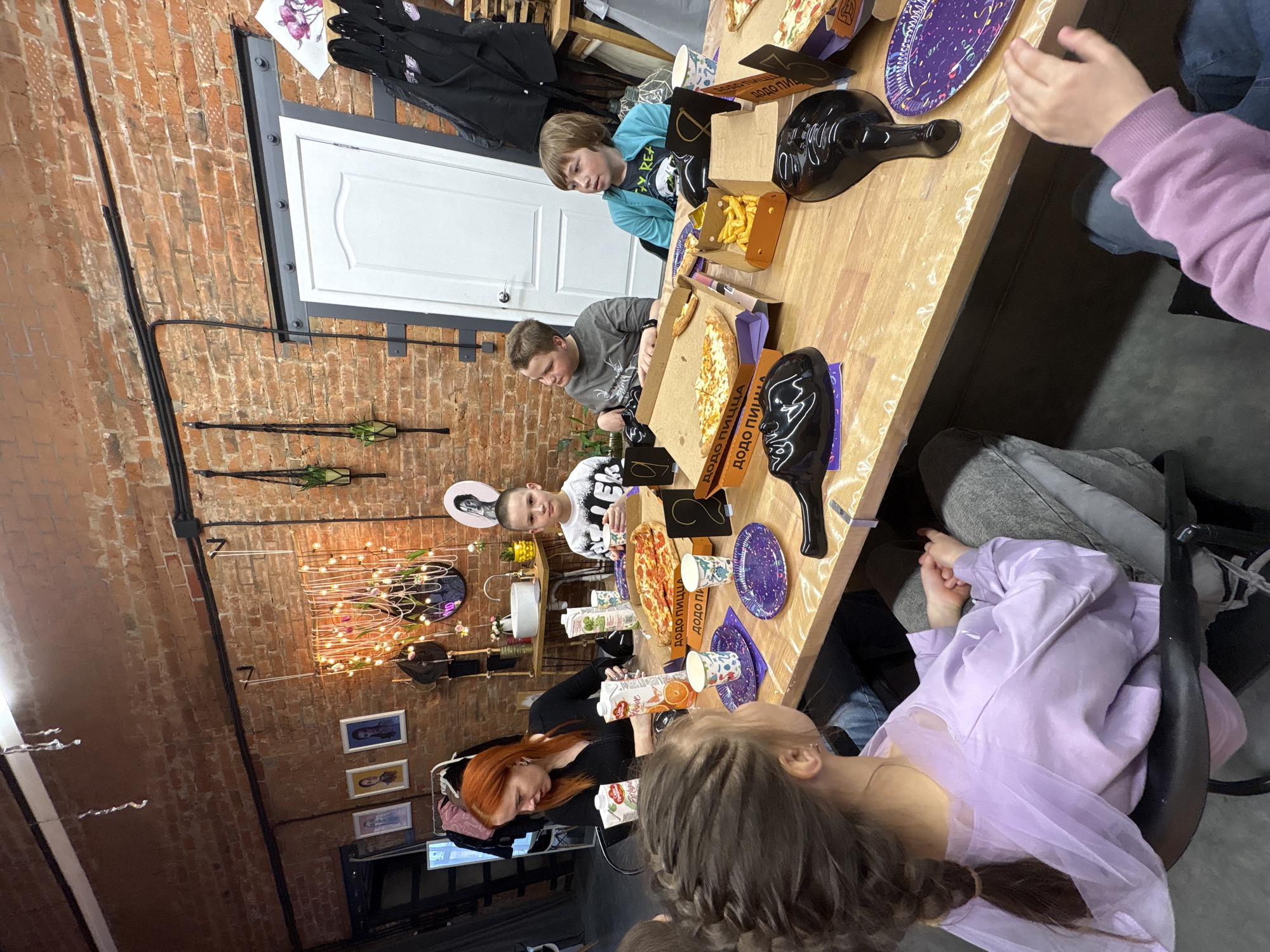 This image captures an indoor gathering, likely a children's party, taking place in Chaikovsky, Russia. Several children of varying ages and two adults are seated around a long, light-colored wooden table.
The room features exposed brick walls, giving it an industrial or loft aesthetic, with visible black electrical conduits running along the brickwork. Decorations on the wall include warm string lights, small green plants in hanging holders, and framed pictures. A white door is visible in the background, above which black coats or bags are hanging.
The central activity involves the group eating and socializing. Multiple open pizza boxes are spread across the table, with some clearly displaying the text "ДОДО ПИЦЦА." Slices of pizza, a container of french fries, and several patterned paper cups and purple party plates are also present. One red-haired adult is seen pouring orange juice from a carton into a cup. Decorative black, shiny, skull-like or mask-shaped items are interspersed among the food on the table.
The children appear casually dressed and engaged in the meal and company. The overall atmosphere is convivial and relaxed, suggesting a celebration or casual get-together. The lighting is warm and artificial, creating an inviting ambiance.