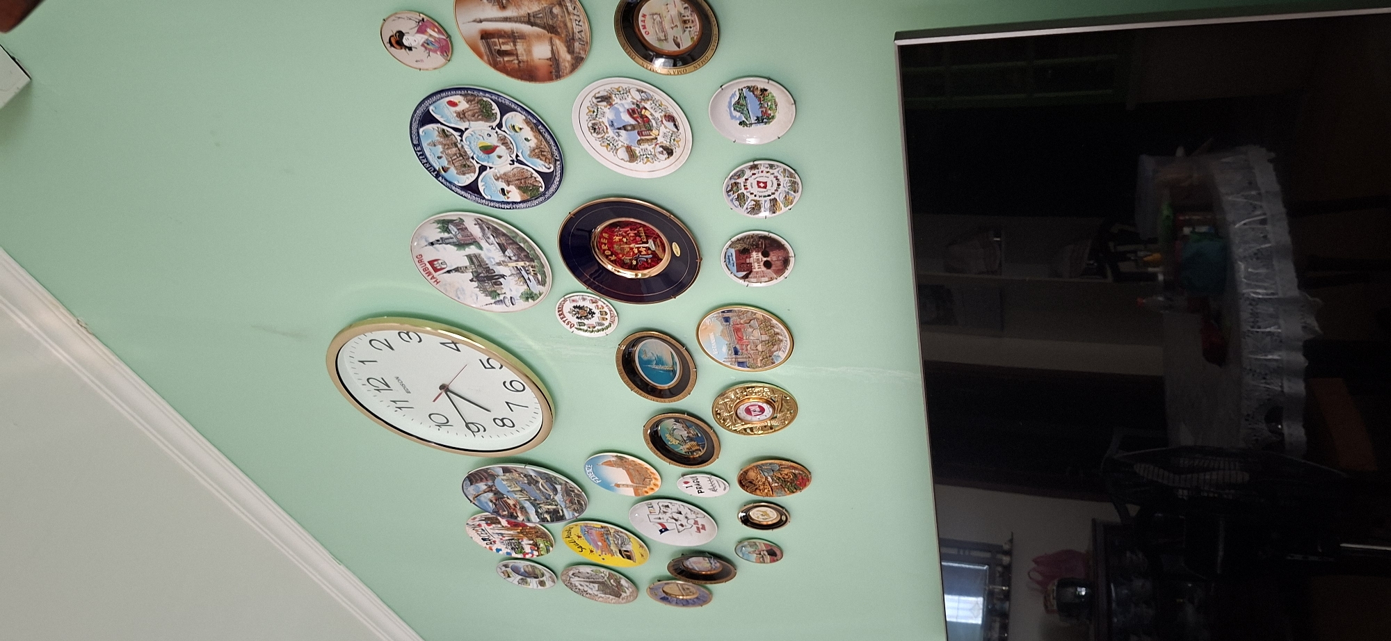 The image shows a wall decorated with numerous decorative plates, each depicting different landmarks or scenes from various cities. There is also a large round clock with Roman numerals on the wall. In the lower portion of the image, a dark television screen is visible. The overall environment appears to be indoors, possibly a living room or dining area, with a suggestion of furniture and a table with items on it in the lower right corner. The wall is painted a light green color. The plates are arranged in a somewhat scattered pattern across the upper part of the wall. No specific location in Surabaya, Indonesia is discernible from the image.