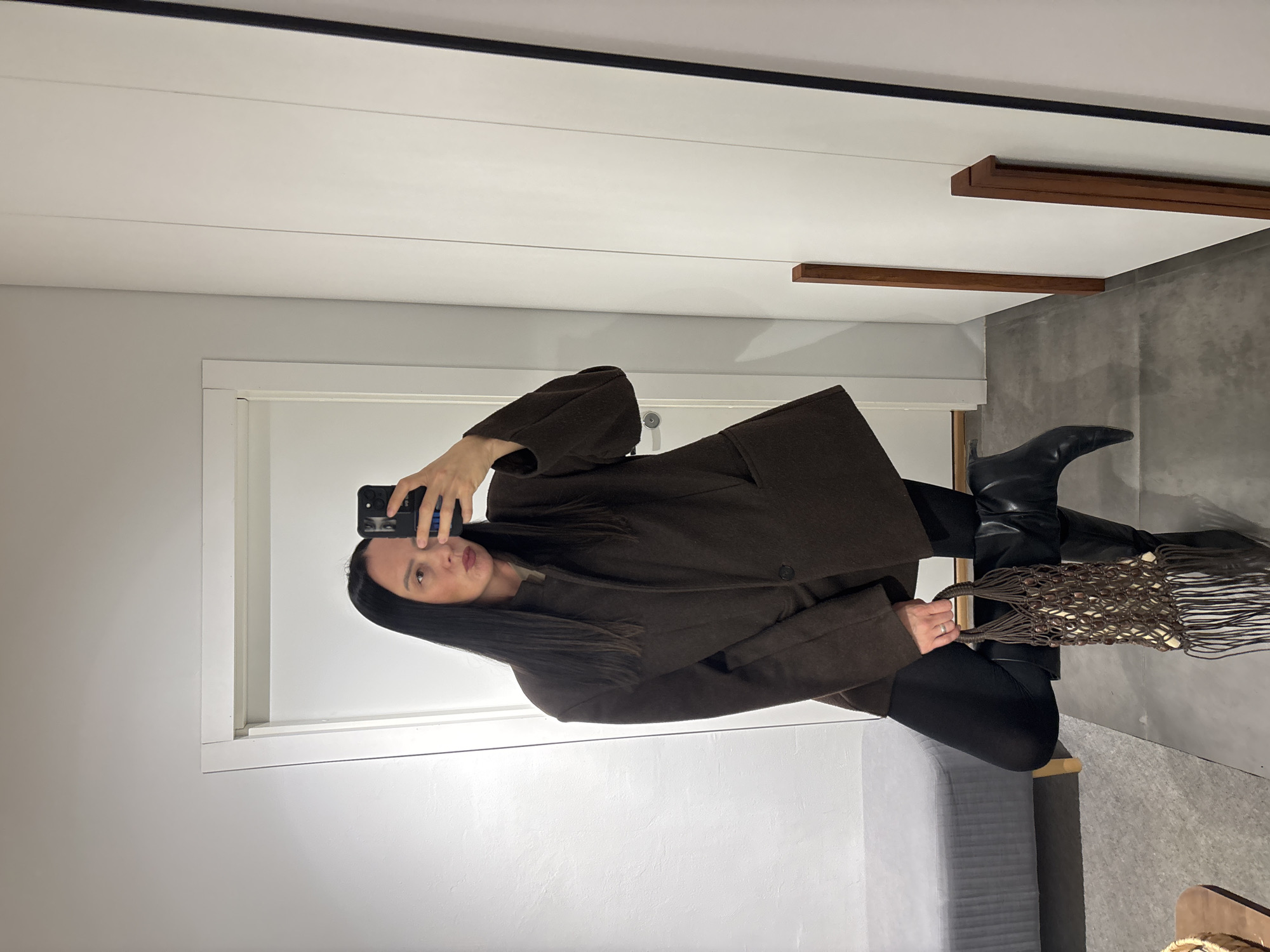 A mirror selfie captures a woman posing indoors, likely within a changing room or a modern interior space in Chaikovsky, Russia. She has long, straight dark hair and is wearing a dark brown, possibly wool, oversized coat or blazer, dark pants or leggings, and black pointed-toe boots. She holds a black smartphone with a unique phone case featuring eyes, using it to take the photograph. In her other hand, she carries a distinctive brown macrame-style string bag, detailed with woven beads and long tassels. A silver-colored ring is visible on her left hand.
The setting, reflected in the large mirror, features bright white walls and a white ceiling with a black linear accent and two decorative horizontal wooden beams. The floor is a dark, possibly concrete or tiled, surface. A portion of a white doorframe is visible, and a grey upholstered bench or seating area is situated behind the woman. Her facial expression is neutral with a slight pout, contributing to a composed and stylish demeanor. The ambient light suggests it is daytime.