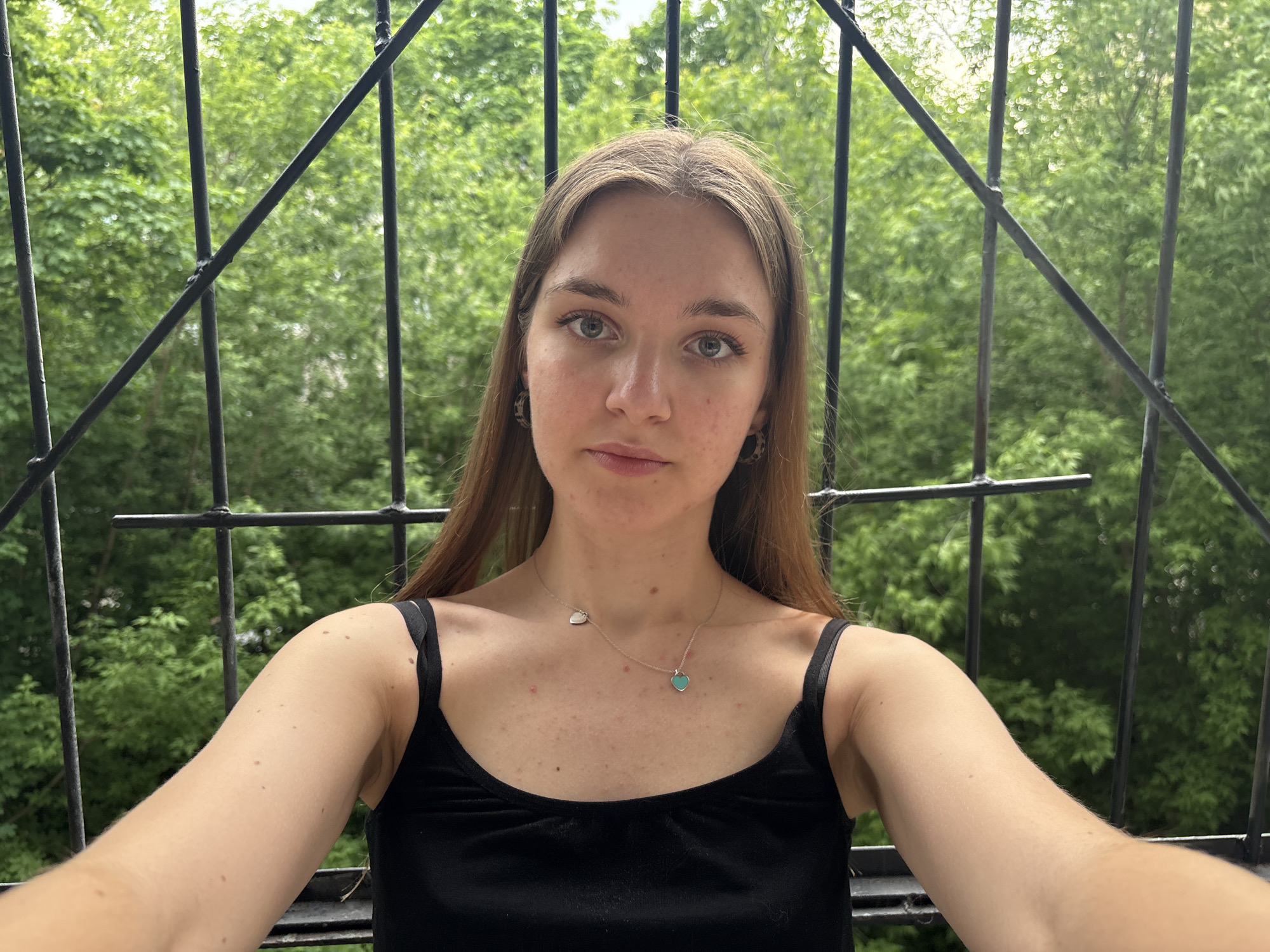 A young woman is centered in the frame, facing the camera. She has long, straight brown hair, fair skin, and light-colored eyes. She wears a black tank top and two necklaces with heart pendants. She appears to be standing on a balcony with a black metal railing, and green trees are visible in the background.
While the image's location within the city of Perm, Russia, cannot be definitively determined from the visual data alone, it is possible that the architecture visible in the background could provide clues if more information about Perm's architectural styles and urban layout were available. Therefore, without further information, it is impossible to confirm the location.