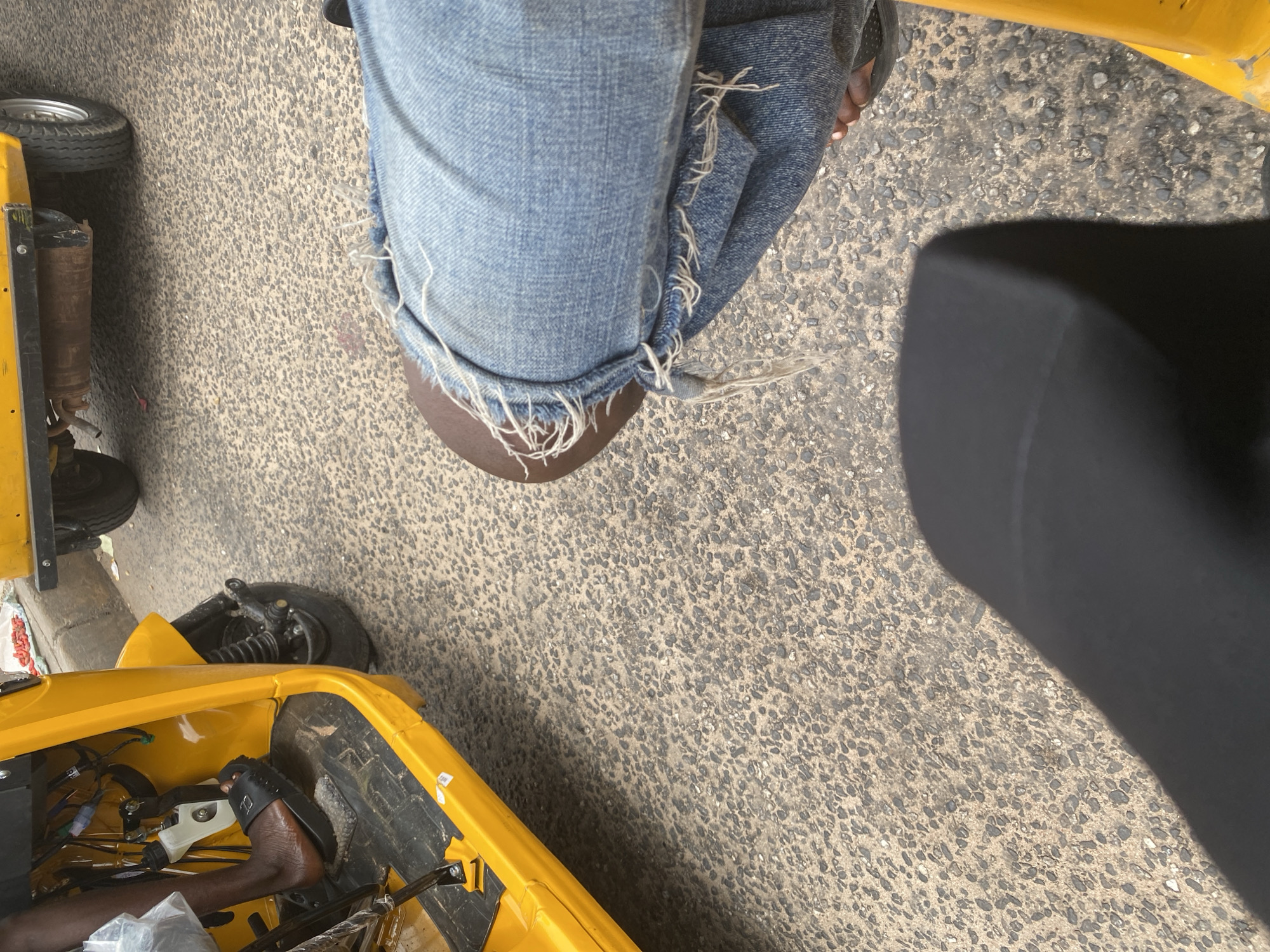 A medium shot captures a person's legs and feet inside a bright yellow three-wheeled vehicle, likely a tuk-tuk, on a paved road. The vehicle's interior is visible, showing the floorboard and some mechanical components. The person is wearing torn denim jeans and black sandals. The road is made of asphalt with visible pebbles and debris. In the background, another three-wheeled vehicle is partially visible, suggesting an urban street scene in Jalingo, Nigeria. The lighting indicates it is daytime.